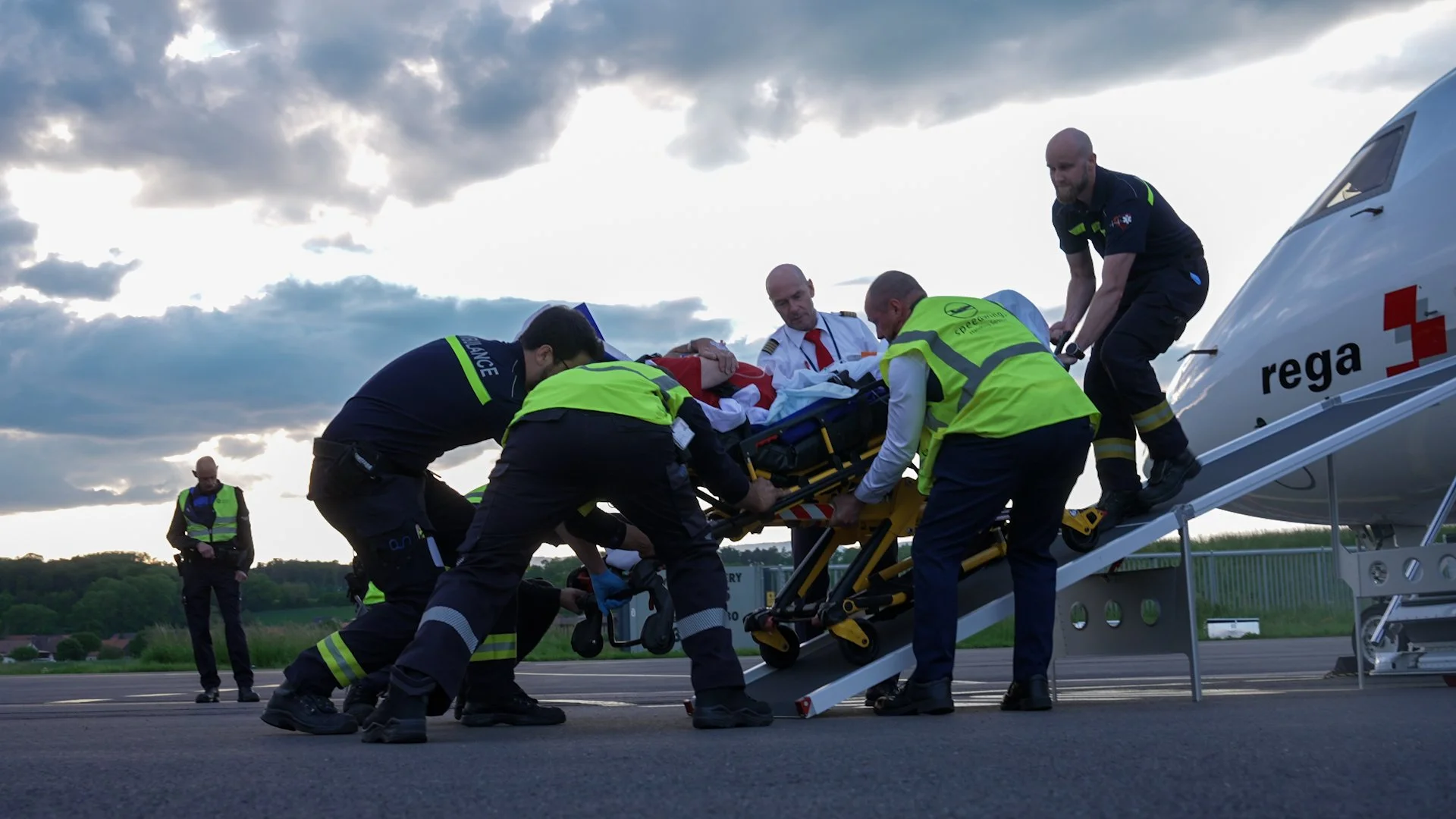 Notärzte und Rettungskräfte transportieren eine verletzte Person auf einer Trage zu einem Ambulanzflugzeug mit angeschlossener Rampe. Es ist Abend oder früher Morgen, bewölkter Himmel im Hintergrund.
