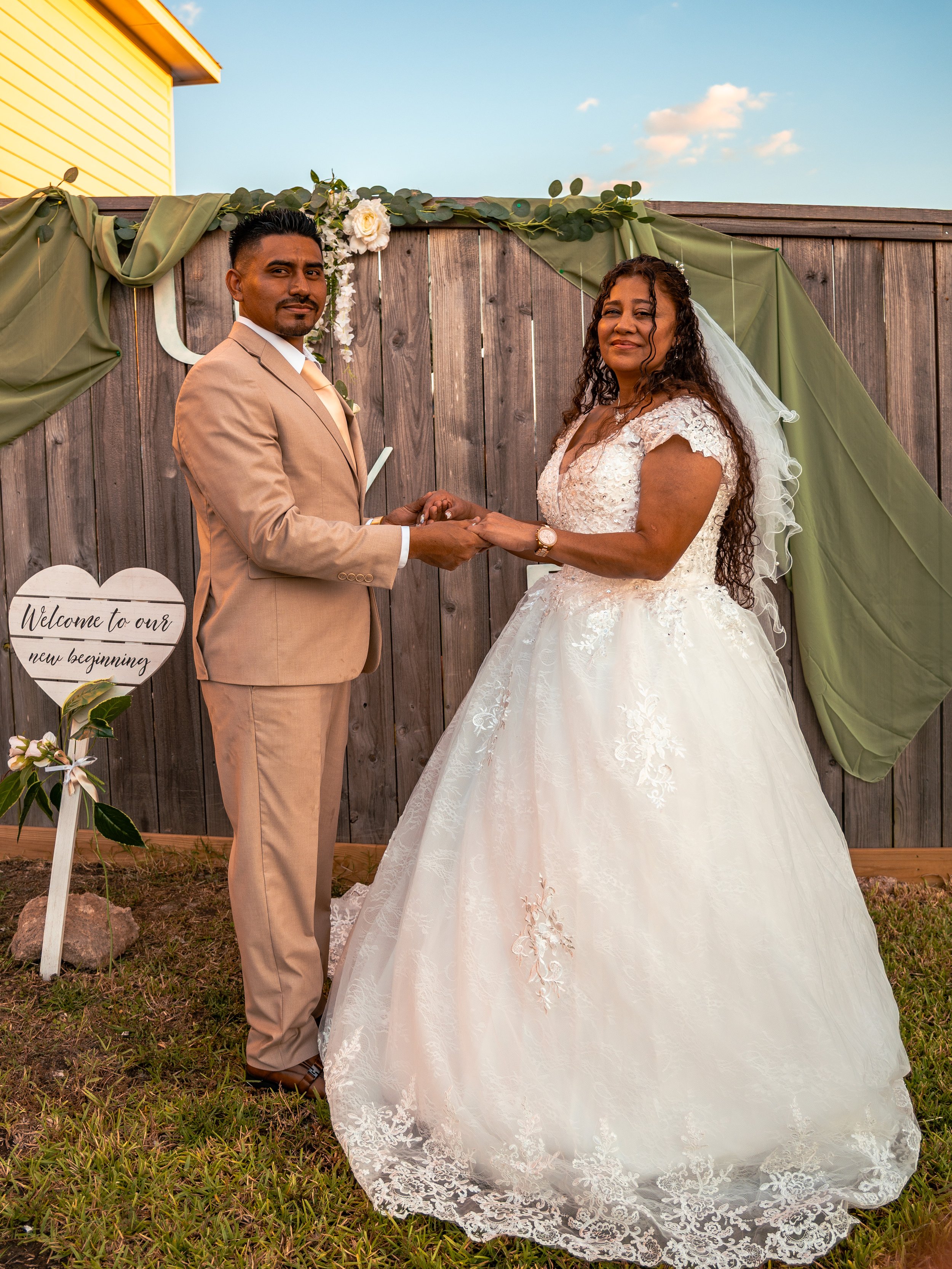 A bride and groom holding hands during their outdoor wedding ceremony, standing in front of a wooden fence decorated with green fabric and white roses, looking at the camera for a photographer in Houston, Texas, to take a picture.