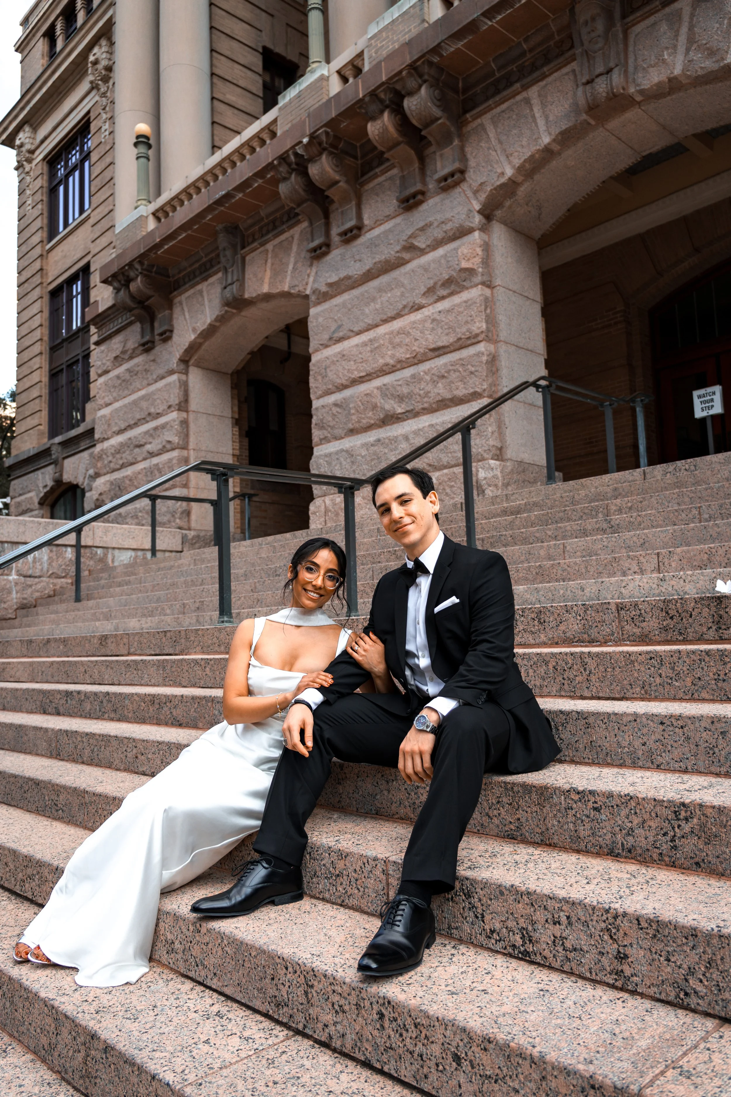 A couple dressed in formal attire sitting on steps outside a historic building, smiling towards a Houston Photographer.
