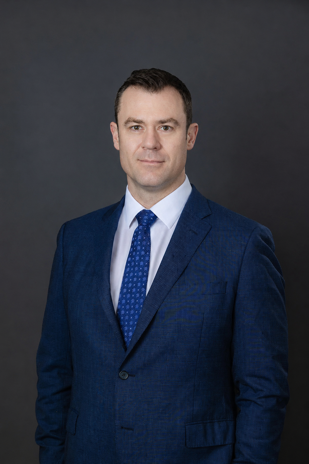 A professional portrait of a man in a blue suit and tie, standing against a dark grey background.