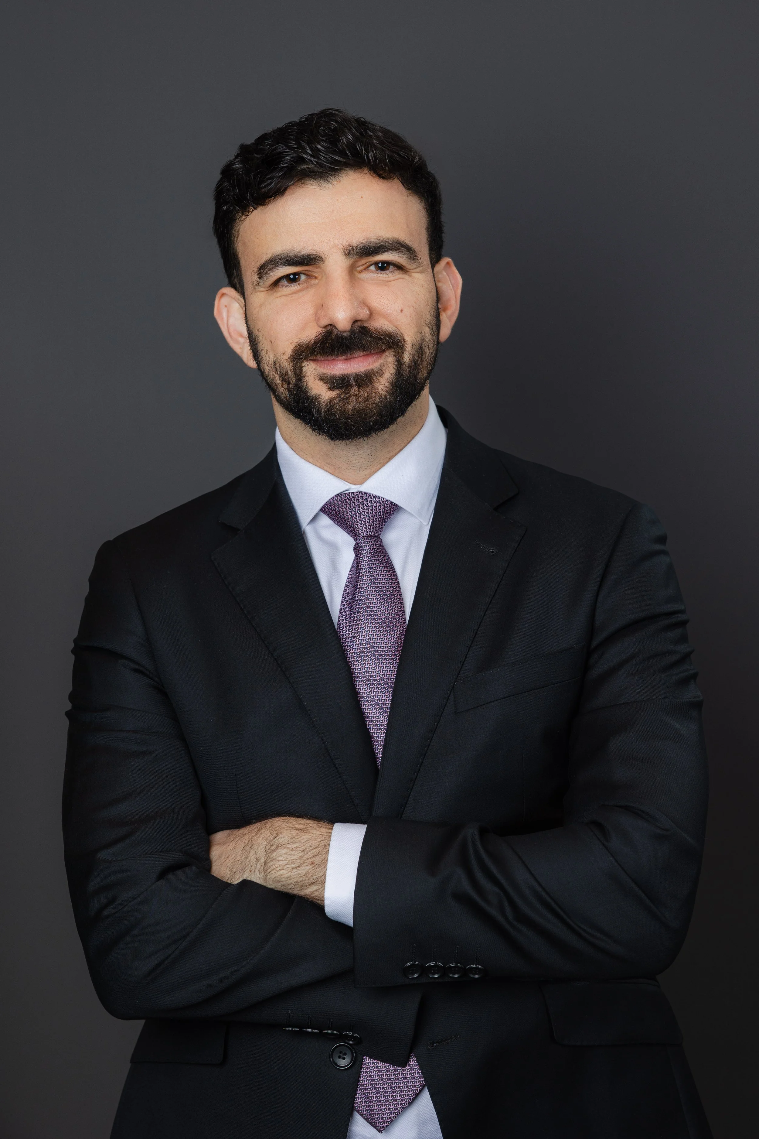 A professional portrait of a man in a black suit, white shirt, and purple tie, standing against a dark gray background.