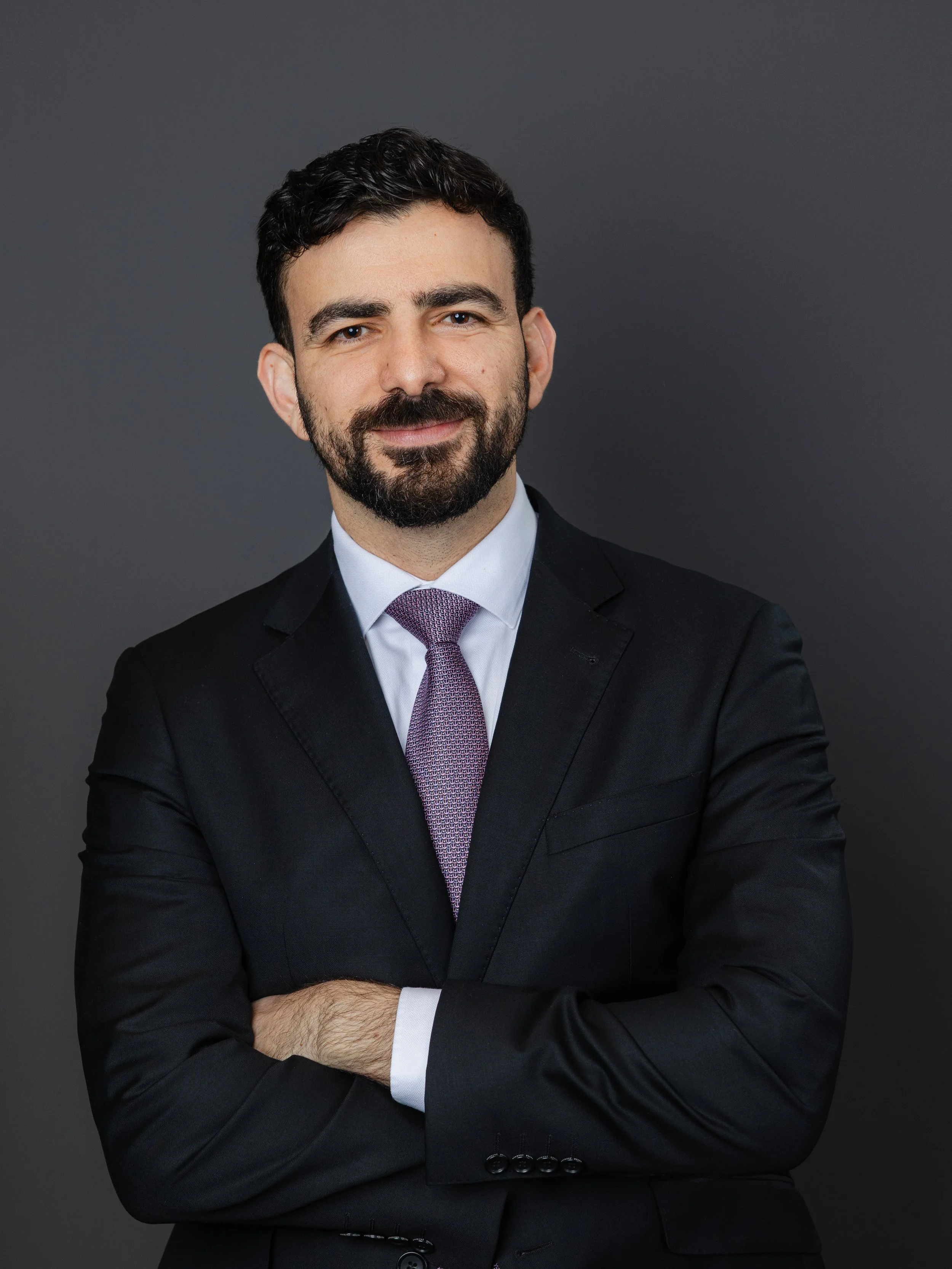 Man with dark hair and beard wearing a black suit, white shirt, and purple tie, standing against a gray background.