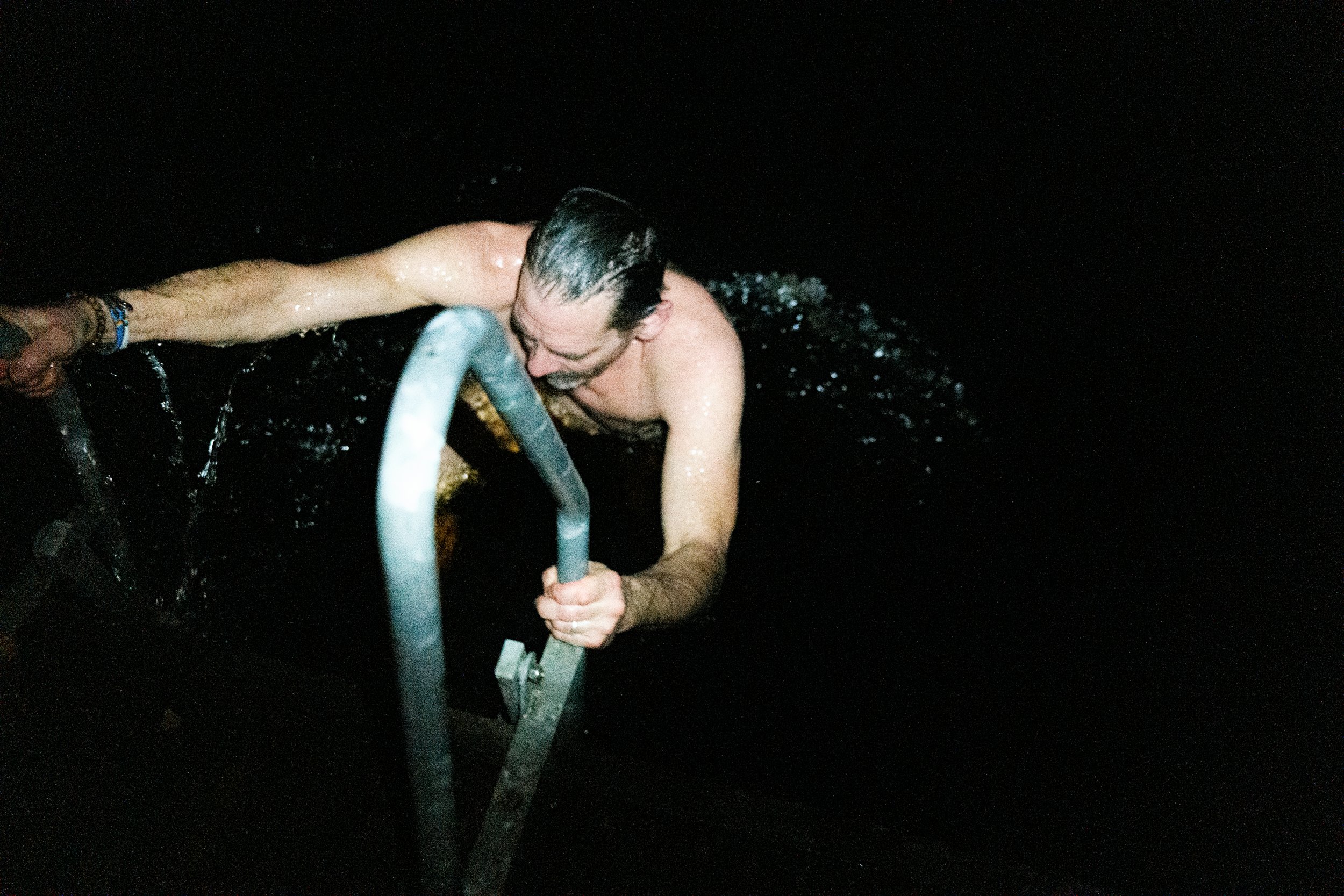 A man with wet hair and a beard climbing a metal ladder out of dark water at night, holding onto the railing with both hands.