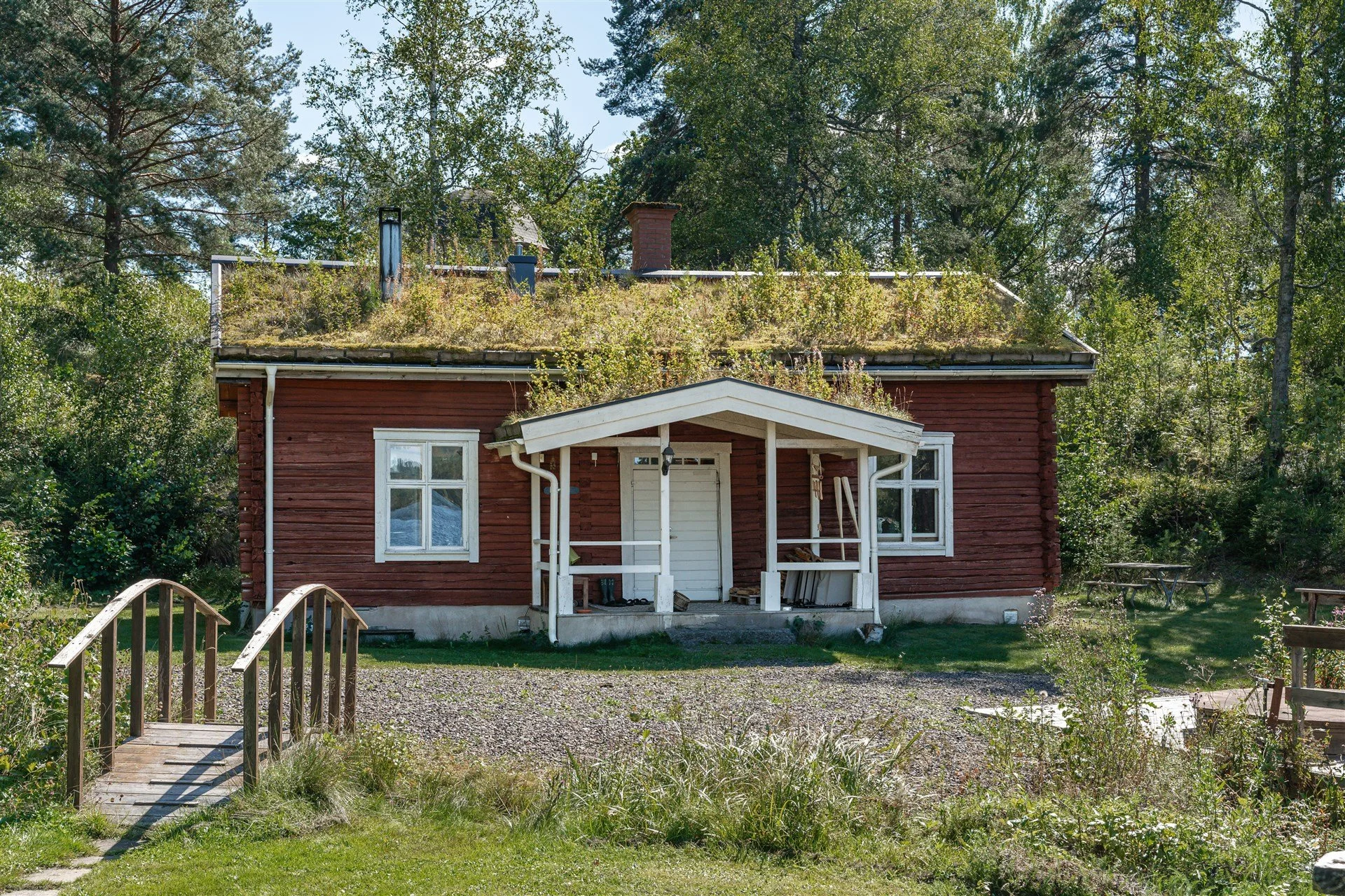 A small red wooden house with a white door and window frames, a front porch with a gabled roof, and a moss-covered green roof. Surrounded by trees and greenery, with a small wooden bridge leading to the house and picnic tables in the yard.