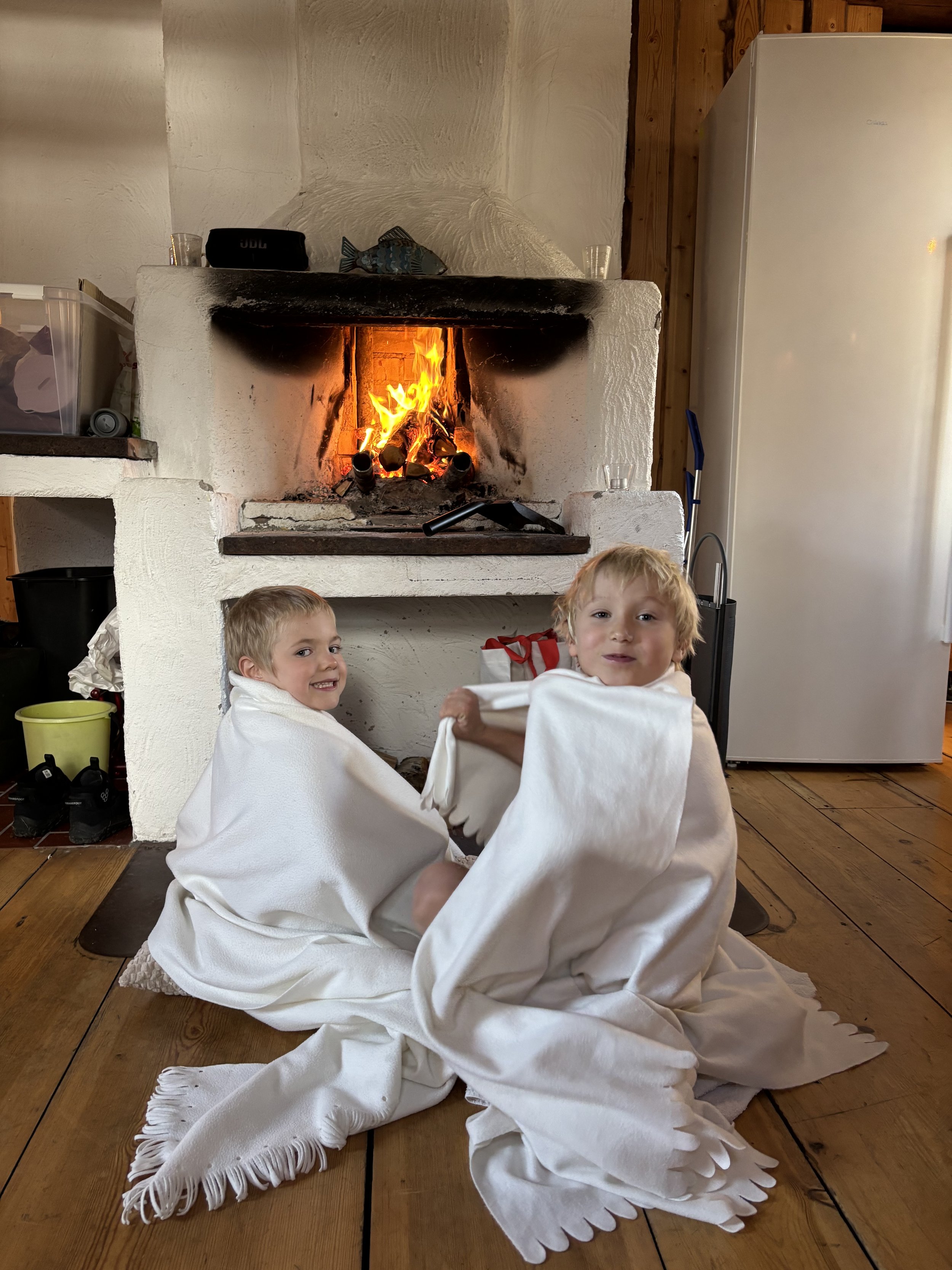 Two young children sitting on the wooden floor near a fireplace with a fire burning inside. They are wrapped in white blankets, smiling and looking at the camera, in a cozy home setting.