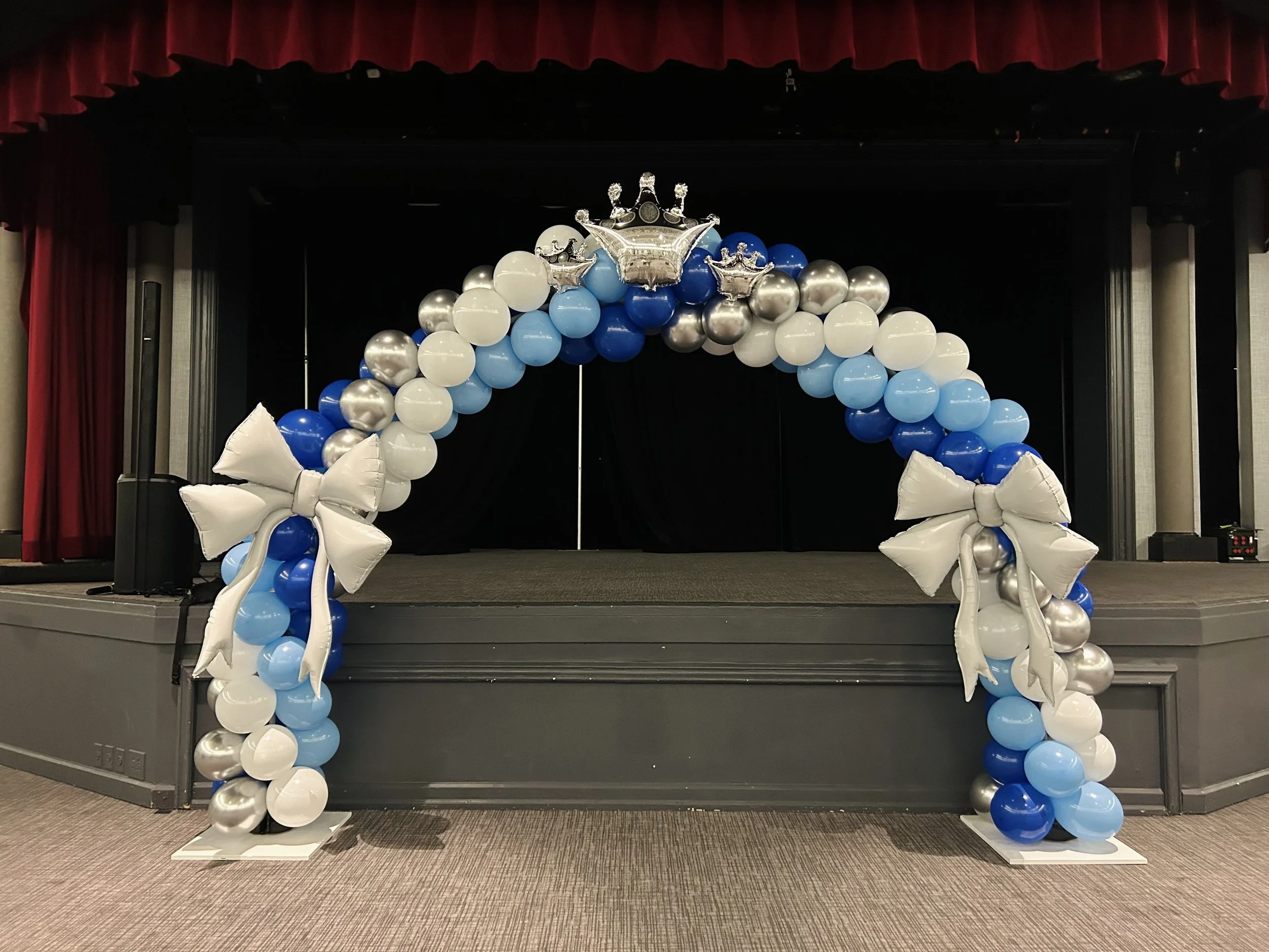 Balloon arch with white, blue, and silver balloons, adorned with large white bows on each side and balloon crowns at the top, set on a stage with red curtains in the background.