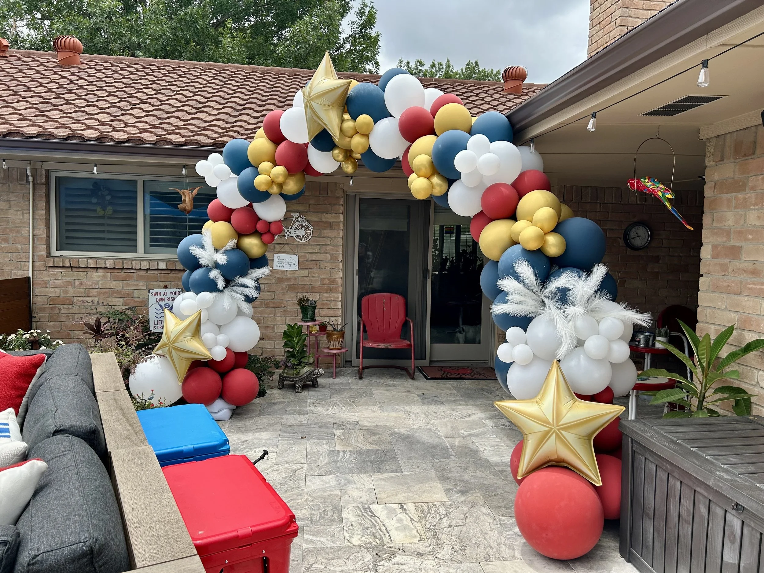 Decorated patio entrance with balloons in red, white, blue, and gold, forming an arch and clusters, with large gold star balloons, white feathers, and a few smaller decorative items hanging around.