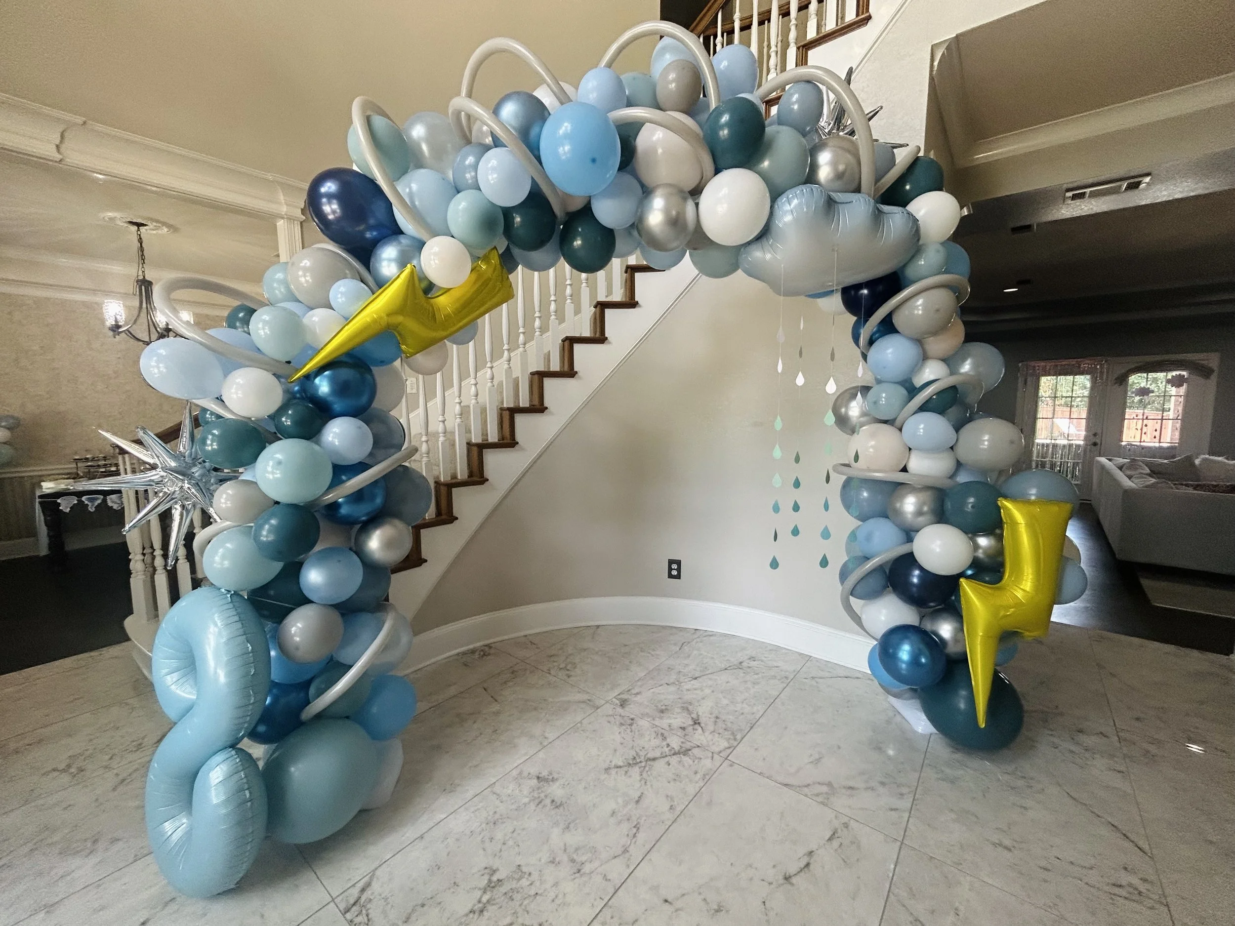 Balloon arch decorated with blue, white, silver, and yellow balloons, featuring star-shaped and number-shaped balloons, set up in a home interior near a staircase.
