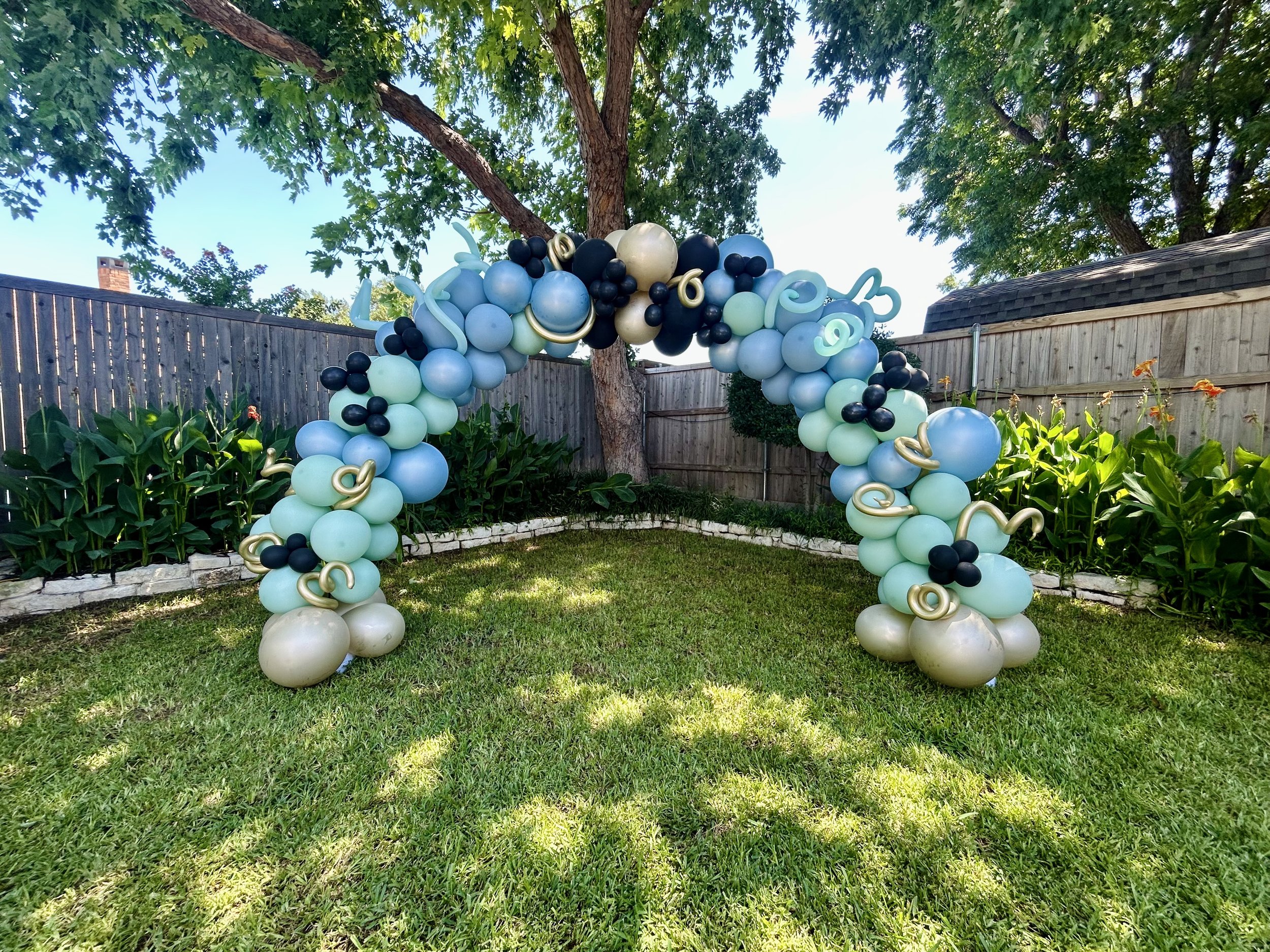 Balloon arch with various shades of blue, black, gold, and cream balloons in a garden with greenery, trees, and a wooden fence.