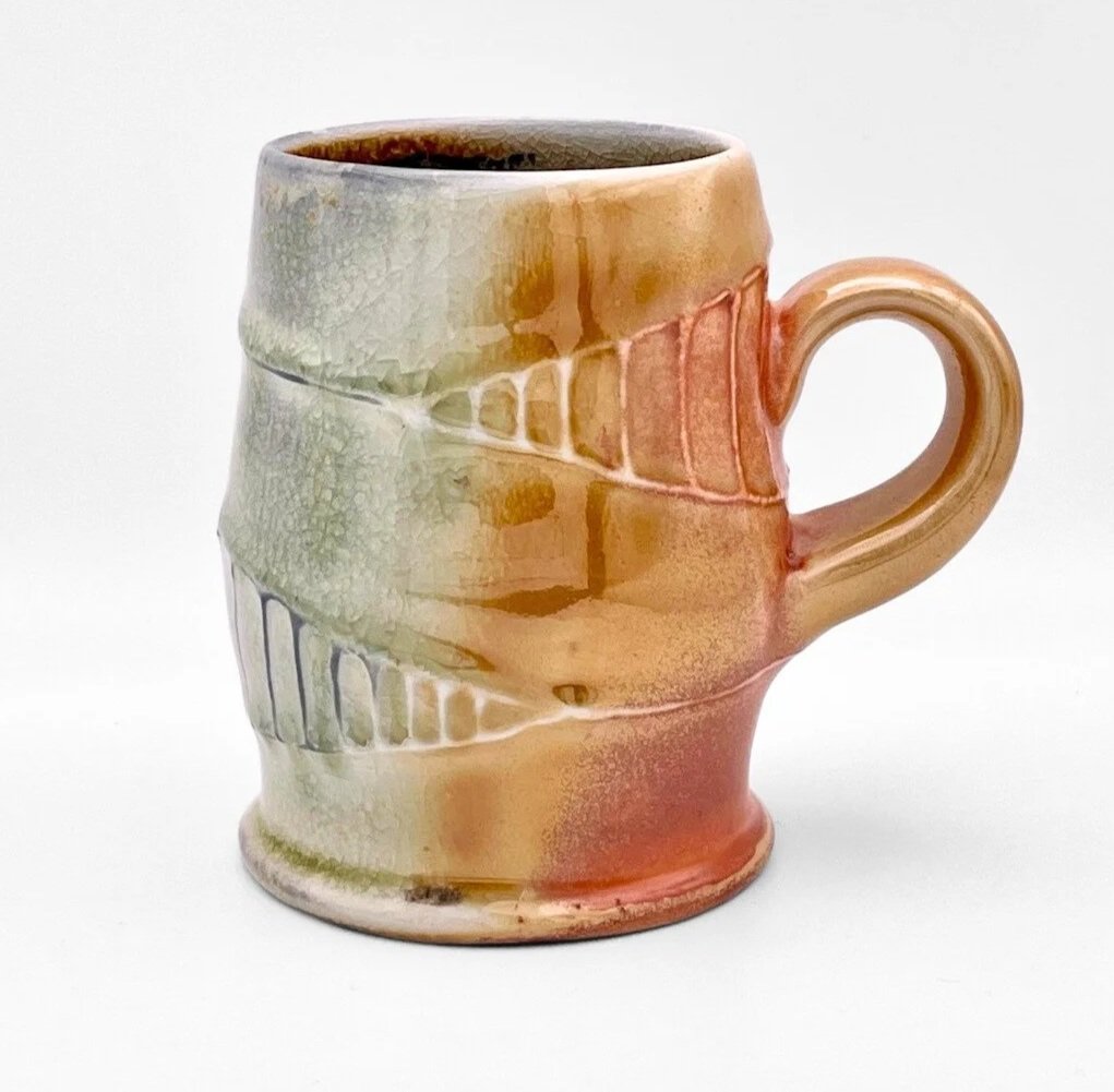 A ceramic mug with an earthy, multicolored glaze, featuring a carved leaf and geometric pattern design, on a white background.