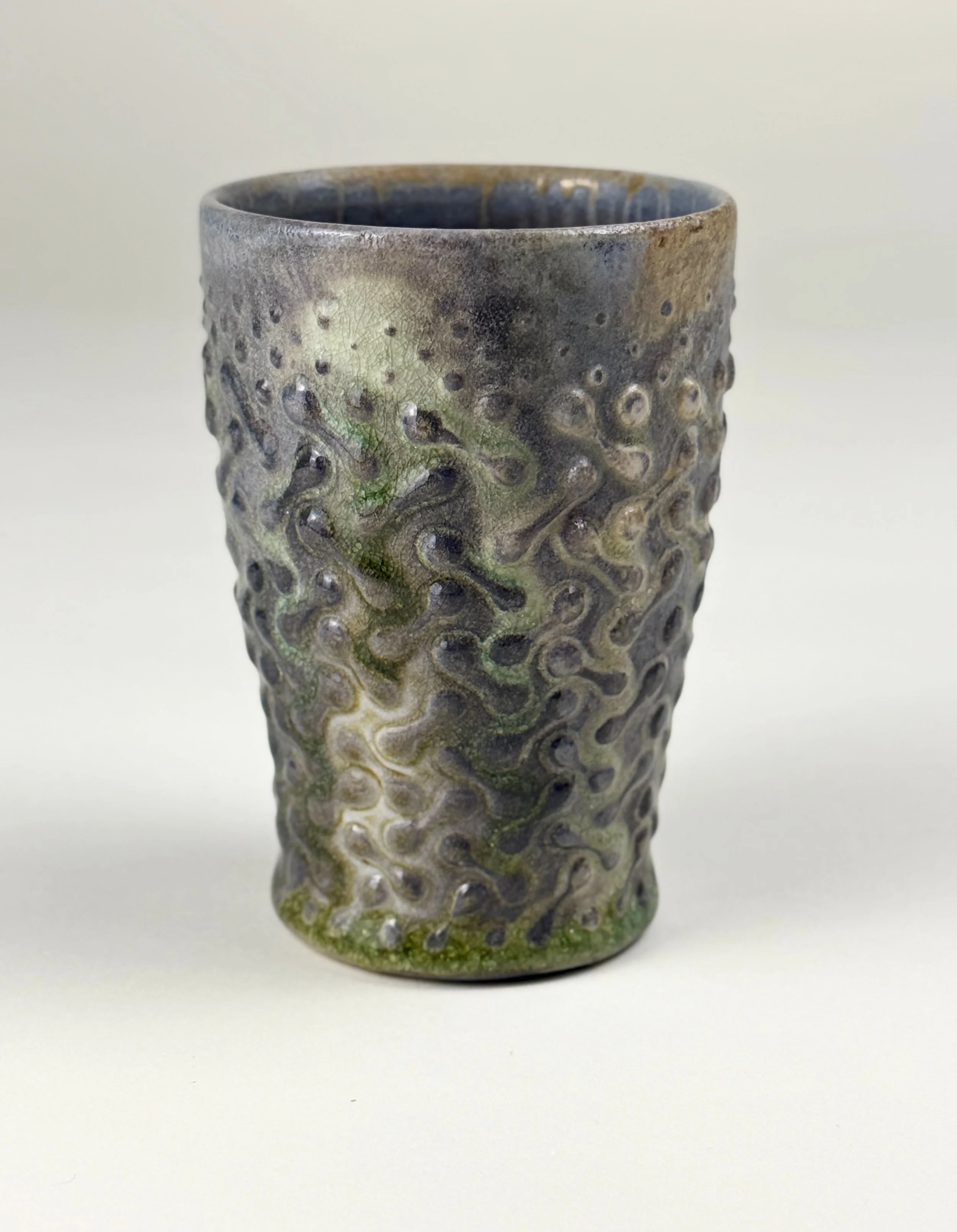 Ceramic cup with a textured, wavy pattern and a gradient glaze of dark and green colors, placed on a plain white background.