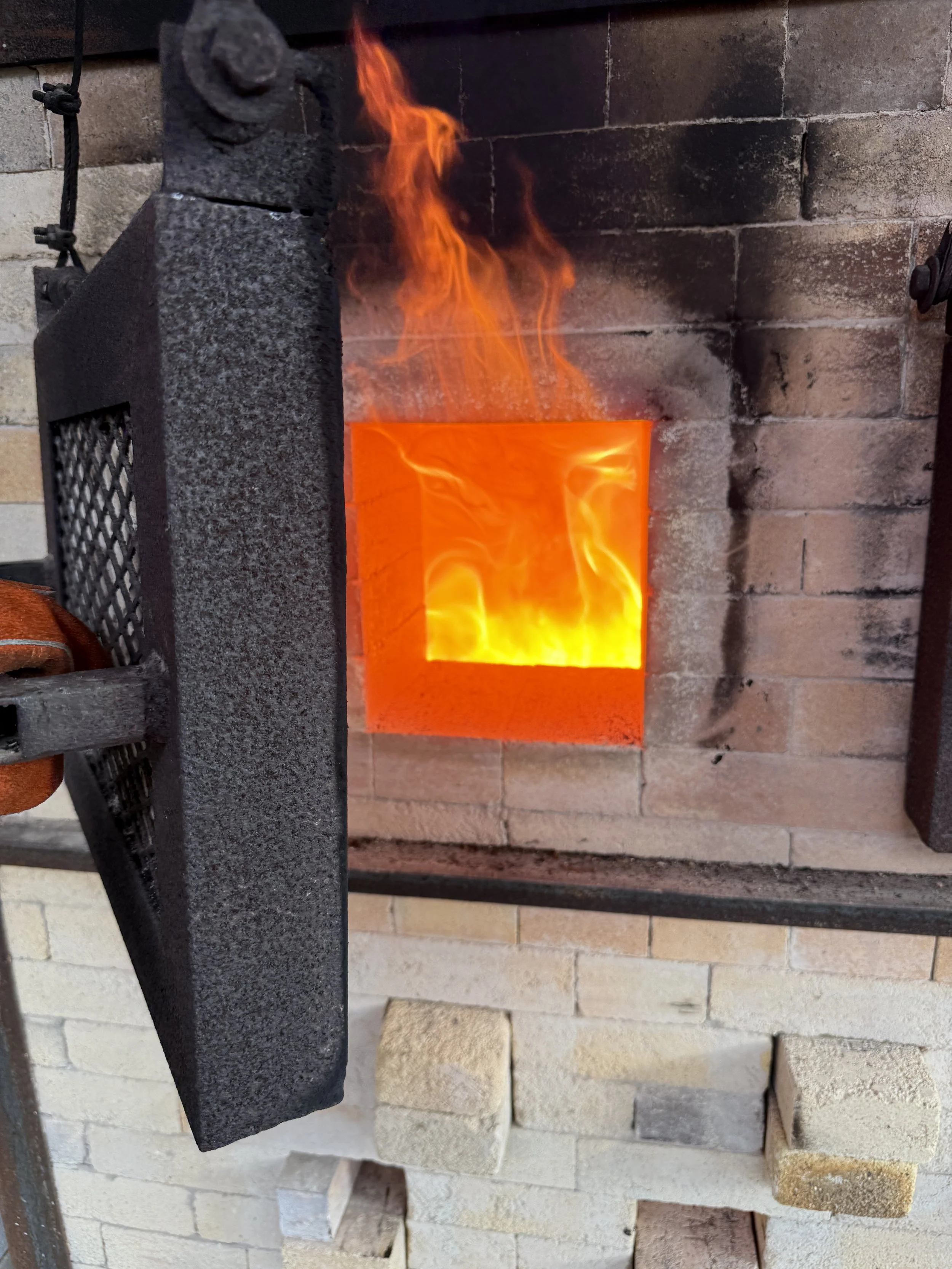 A forge fire with bright orange flames inside a brick-lined hearth, with a metal door or shield partially pulled back.