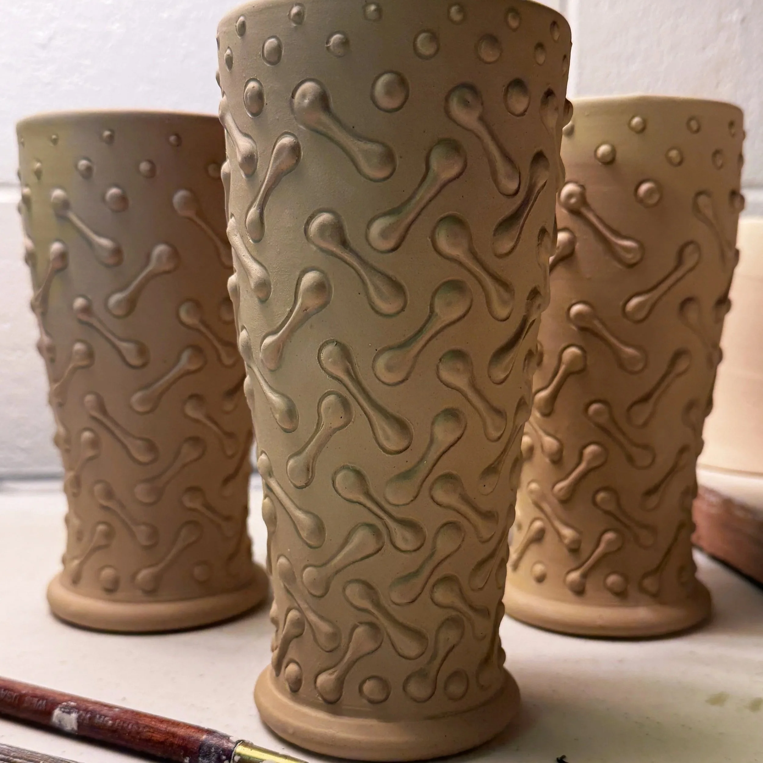 Three tall ceramic vases with a bumpy bone pattern design, placed on a white surface with a paintbrush in the foreground.