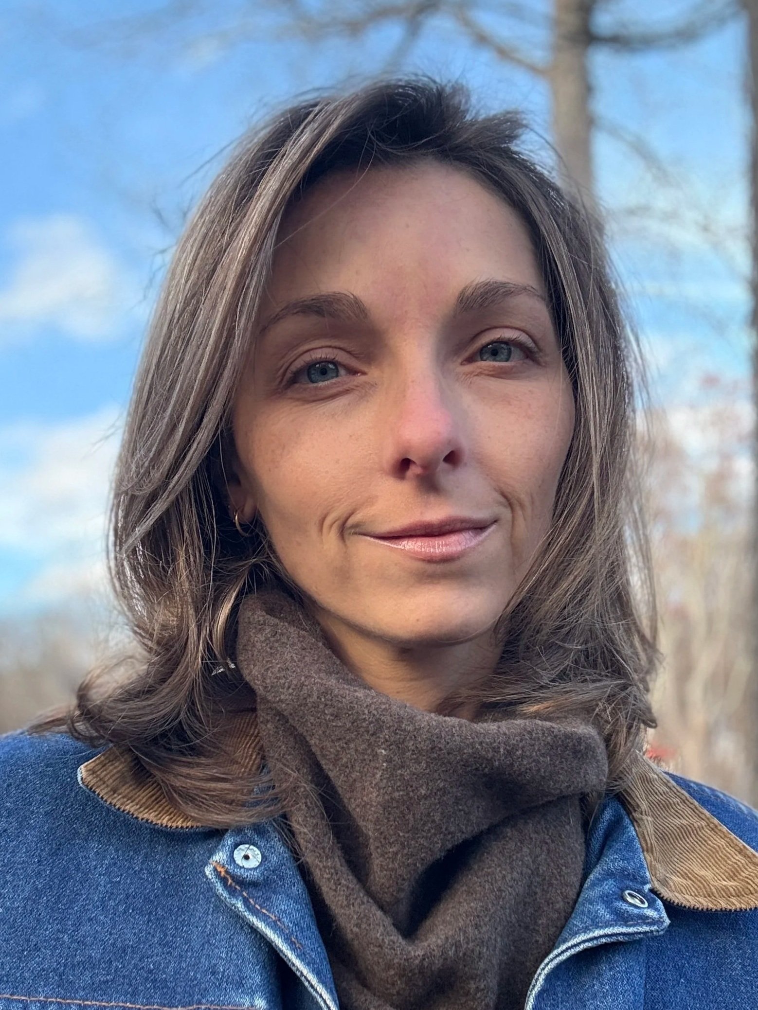 A woman with shoulder-length brown hair, light skin, and blue eyes, smiling outdoors against a background of a blue sky with some clouds and trees.