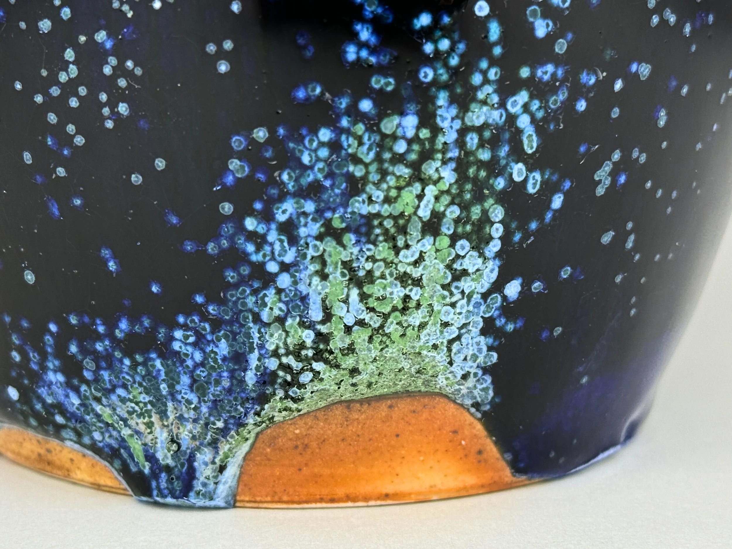 Close-up of a ceramic or pottery surface with a dark background, featuring a vibrant, textured green and blue pattern with small circular and bubble-like details, and a brownish-orange area at the bottom.
