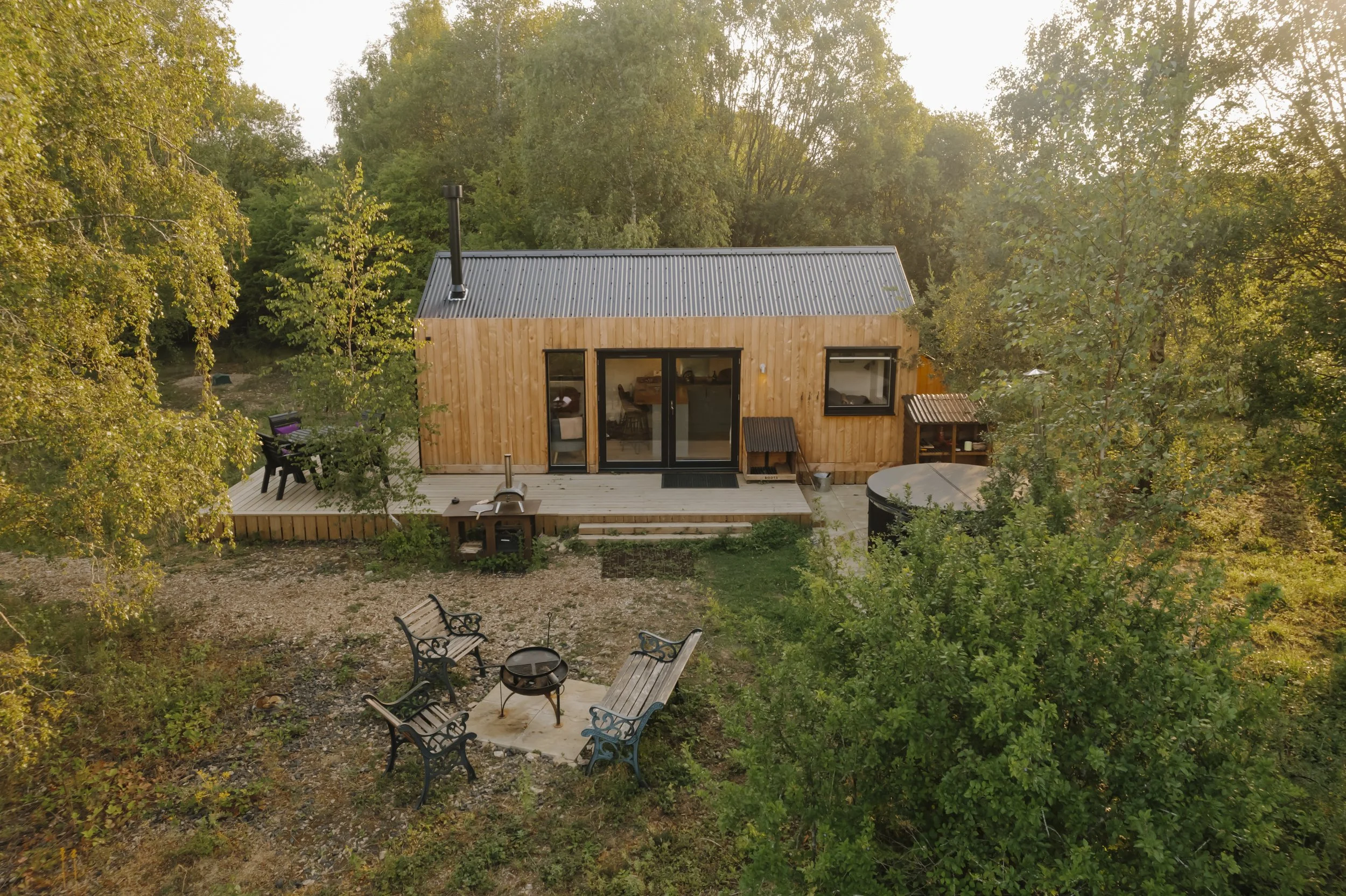 A small wooden cabin with a metal roof surrounded by trees, featuring a deck with outdoor furniture and a grassy yard with chairs and a fire pit.