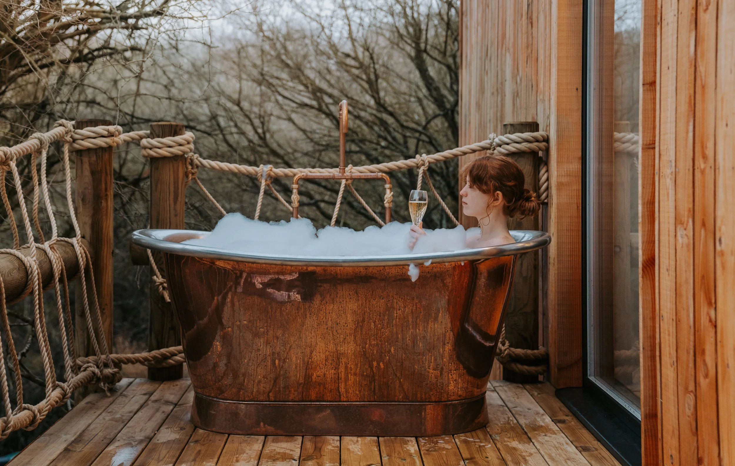 A woman with red hair relaxing in a copper bathtub filled with bubbles on a wooden deck, holding a glass of champagne, surrounded by a wooden railing with rope detailing, overlooking trees.