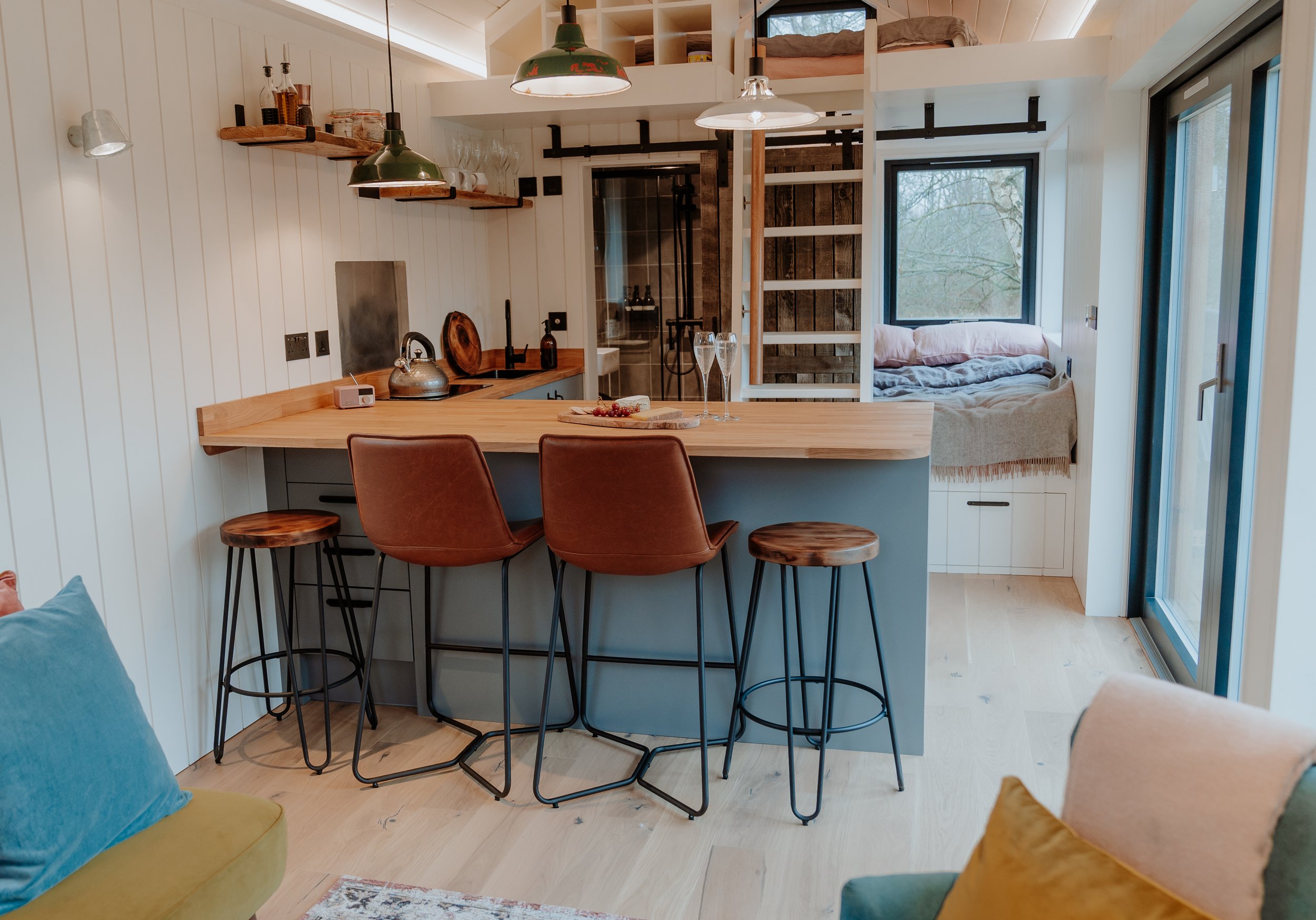Modern kitchen with wooden bar counter, brown bar stools, white wooden wall panels, open shelving with glasses and bottles, small sitting nook with bedding, large window showing outdoor trees, sliding glass door.