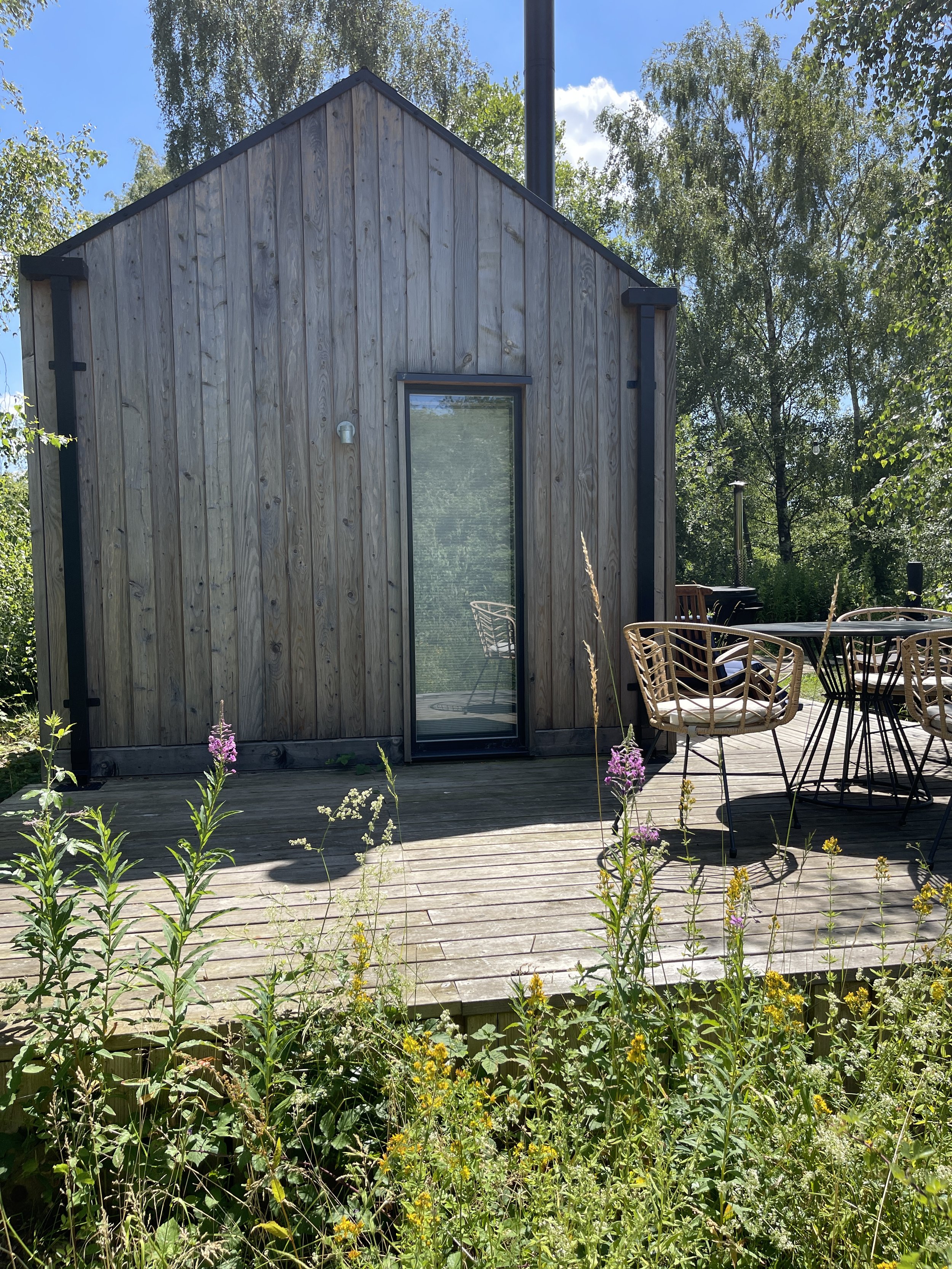 A small wooden cabin with vertical siding, a glass door, and an outdoor patio with chairs and tables, surrounded by wildflowers and greenery under a blue sky.