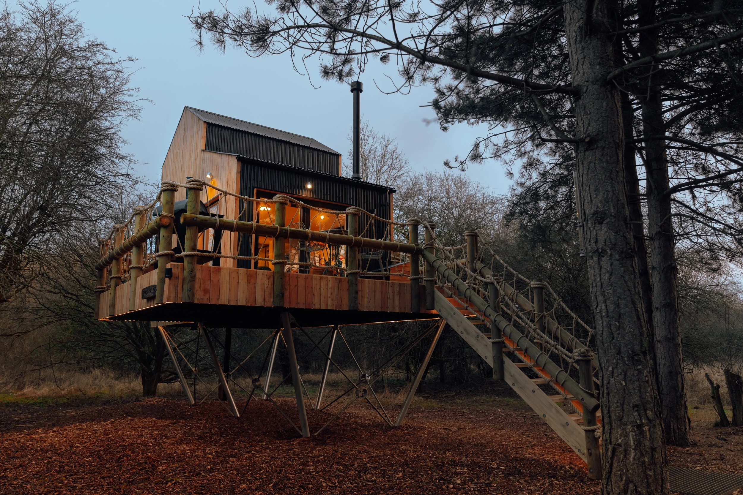 Treehouse built among tall trees, illuminated with string lights, featuring wooden and black metal exterior, with a wooden staircase leading up to it, located in a wooded area during dusk or evening.