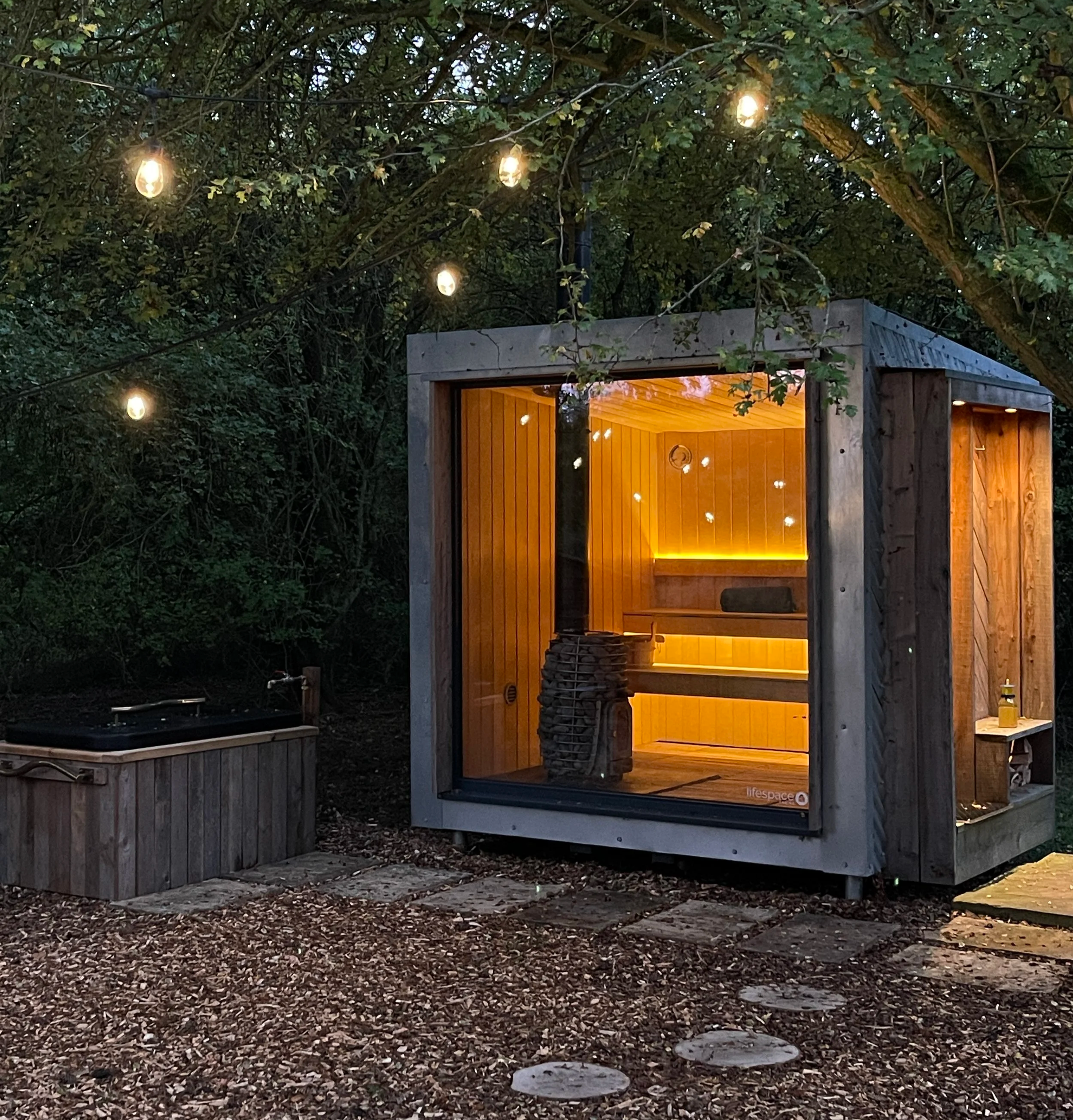 A modern wooden sauna with warm interior lighting in a forest setting, illuminated string lights above, and a small outdoor plunge pool nearby.