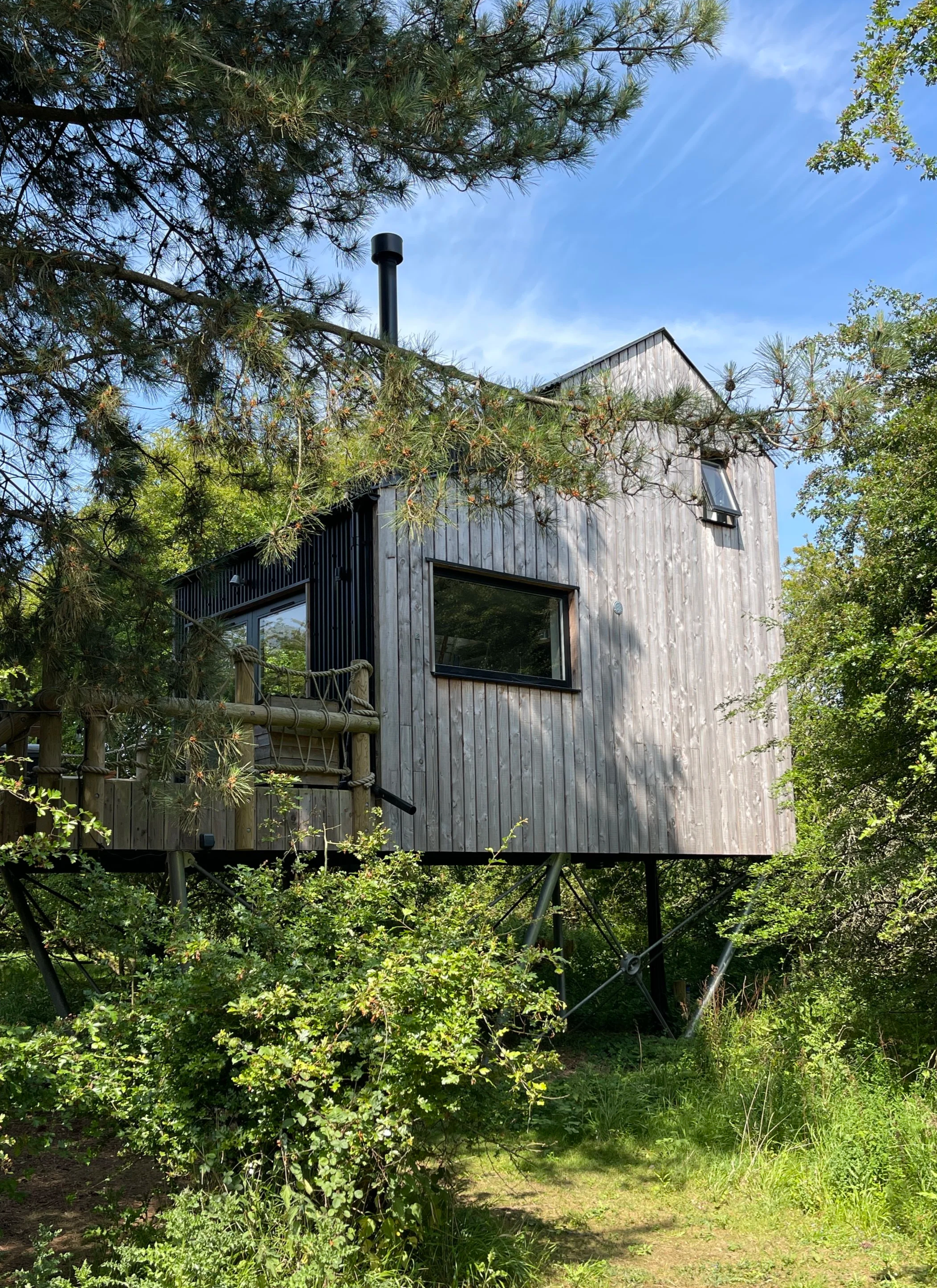 A small modern treehouse elevated on stilts among trees, with a wooden exterior, large windows, and a small balcony, under a partly cloudy blue sky.