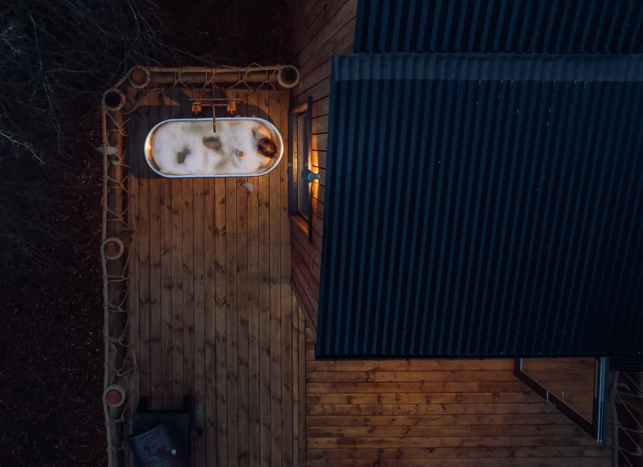 A person soaking in an outdoor bathtub on a wooden deck at night, illuminated by a small light fixture.
