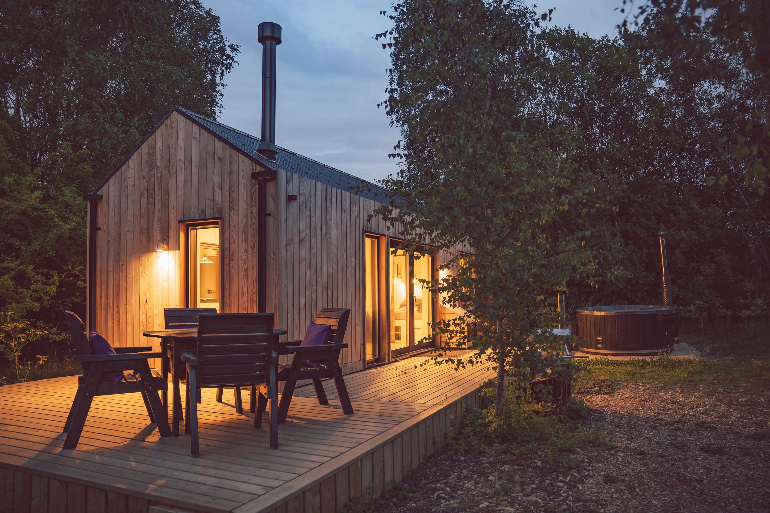 A wooden cabin with lit windows on a deck outdoors at dusk, surrounded by trees, with an outdoor hot tub beside it.
