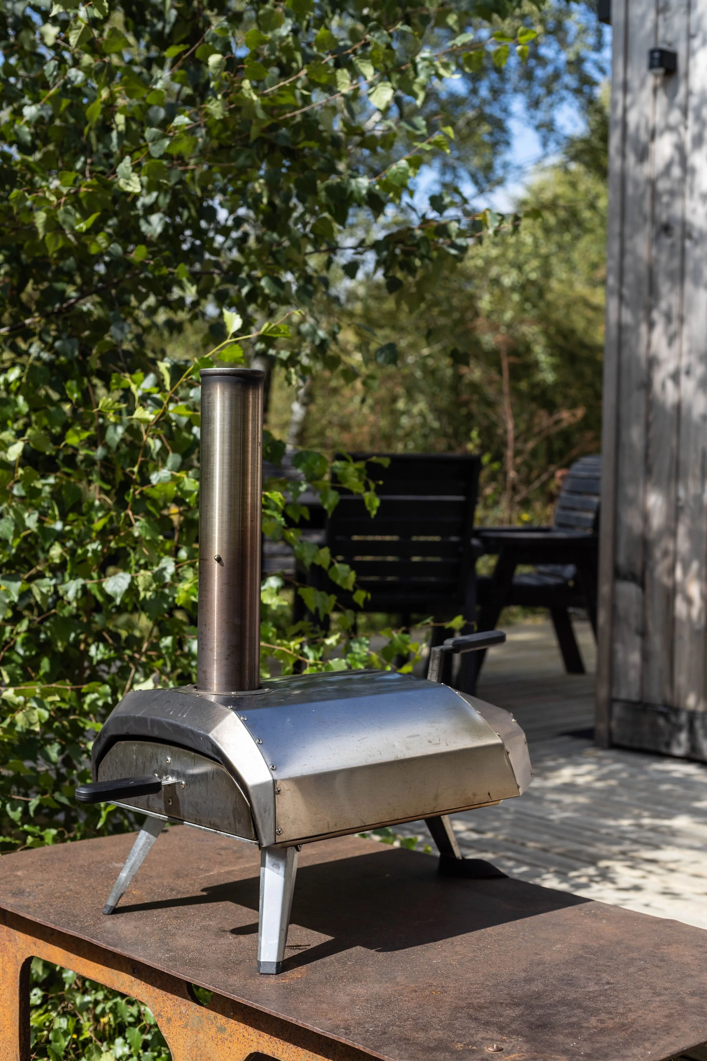A stainless steel portable outdoor pizza oven with black handles and short legs sitting on a rust-colored table in a backyard patio. Green leafy bushes, a wooden deck, and outdoor chairs are visible in the background.
