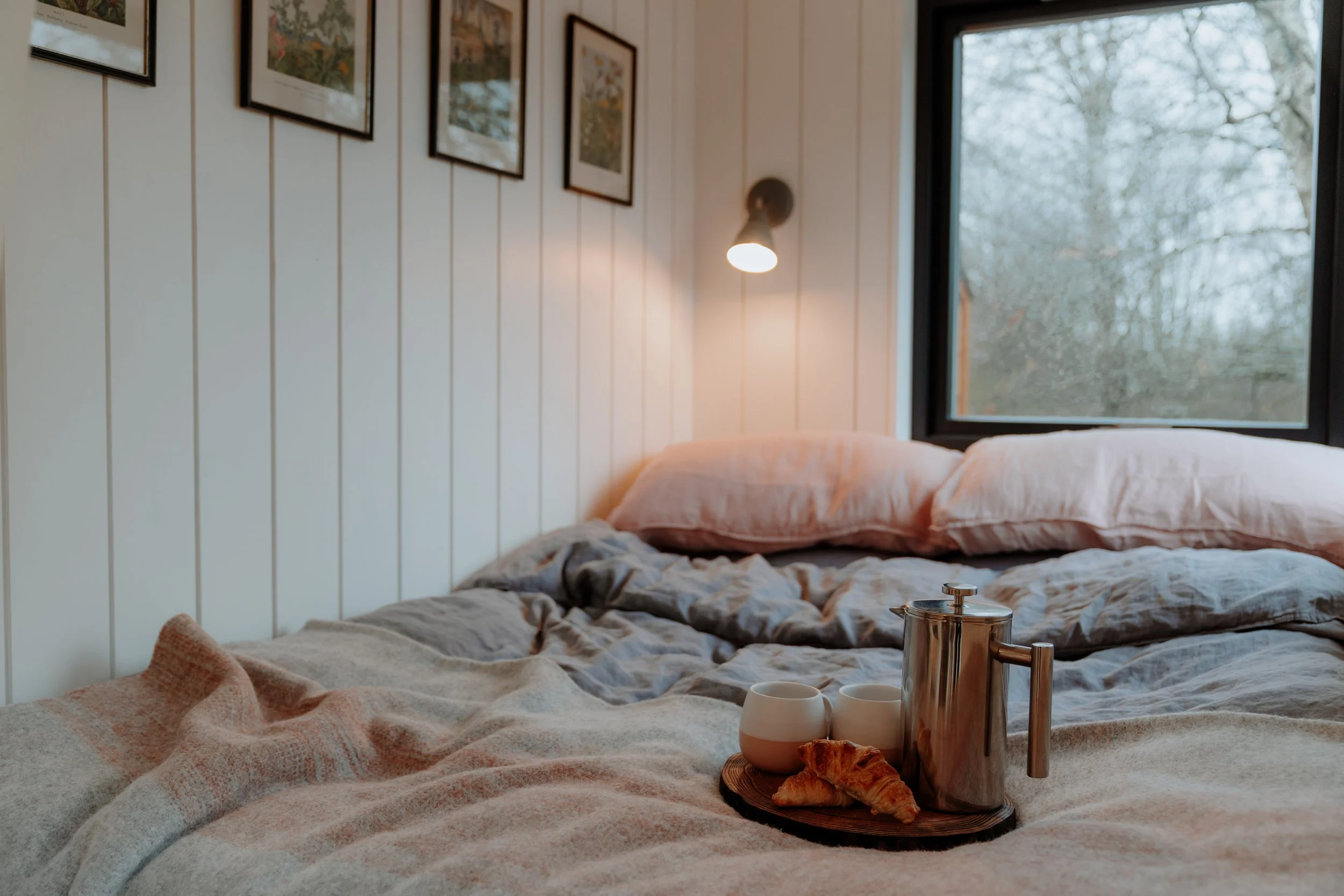 Cozy bedroom with unmade bed, pink pillows, and a tray holding two cups, a croissant, and a stainless steel French press, with a window showing trees outside.