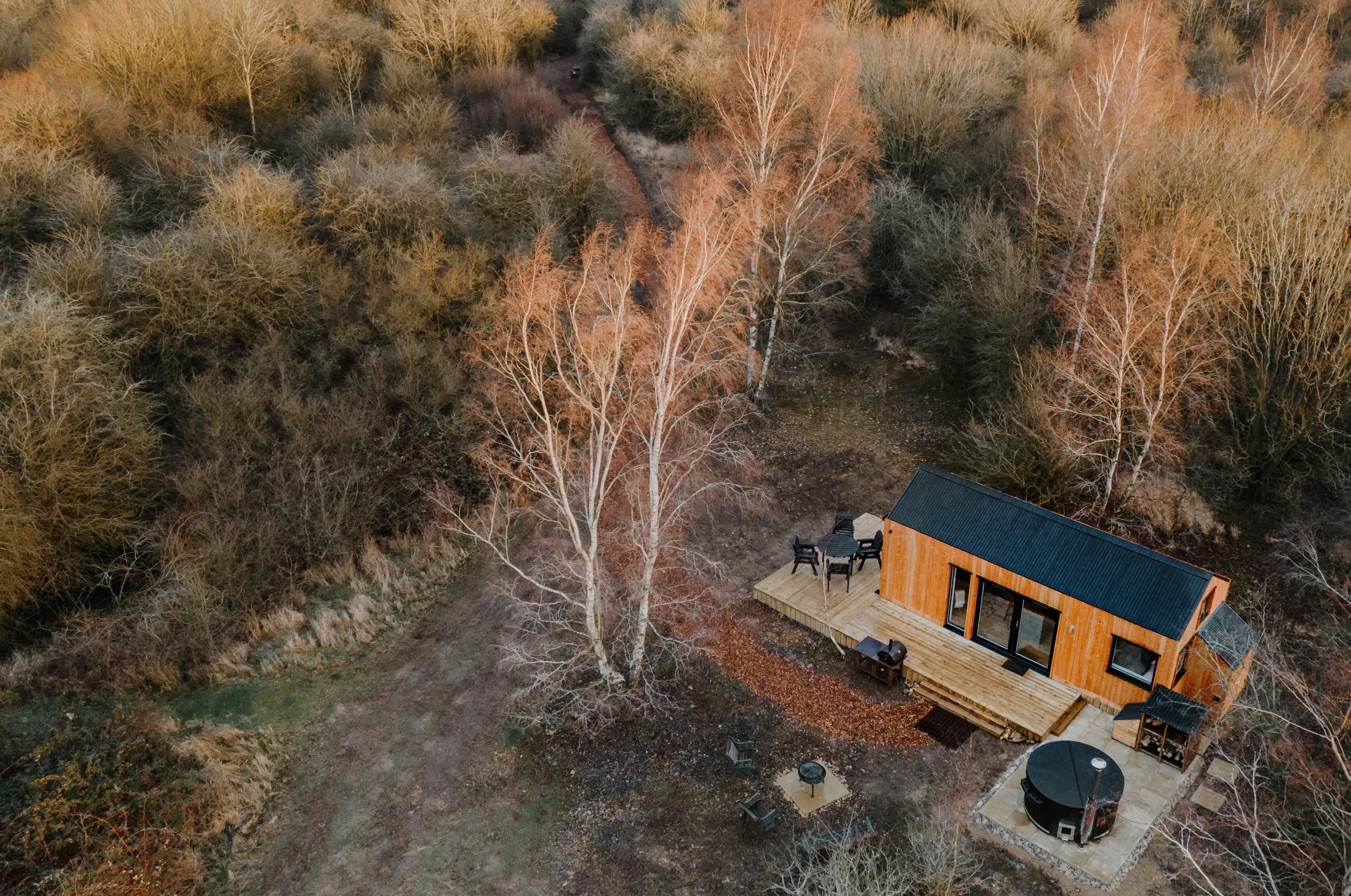 A small wooden cabin with a black roof situated in a wooded area with leafless trees, a deck with outdoor furniture, and a round hot tub nearby, captured from an aerial view during fall or winter.