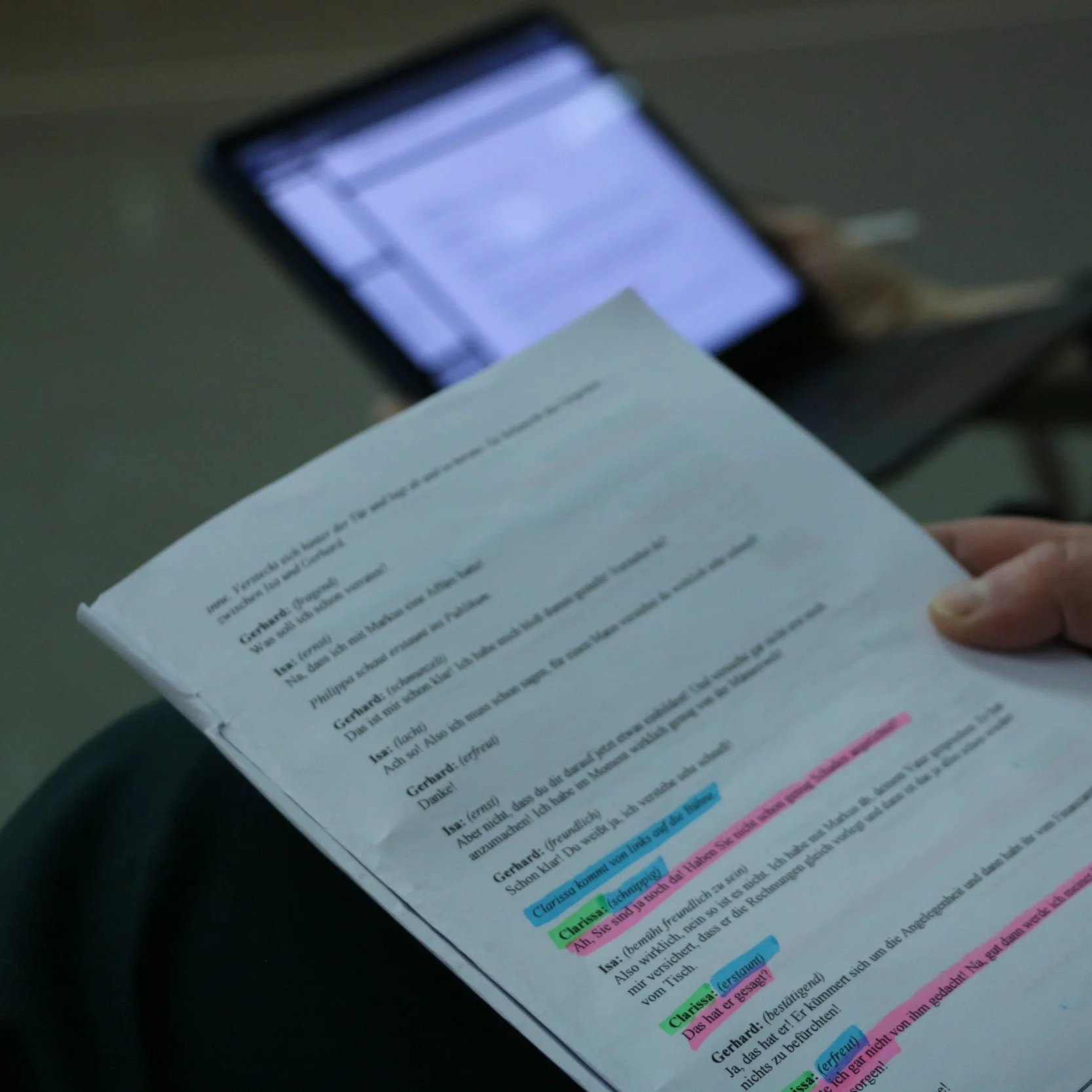 A person holding a printed script or screenplay with highlighted lines and annotations. In the background, there is a blurred laptop screen displaying a document.