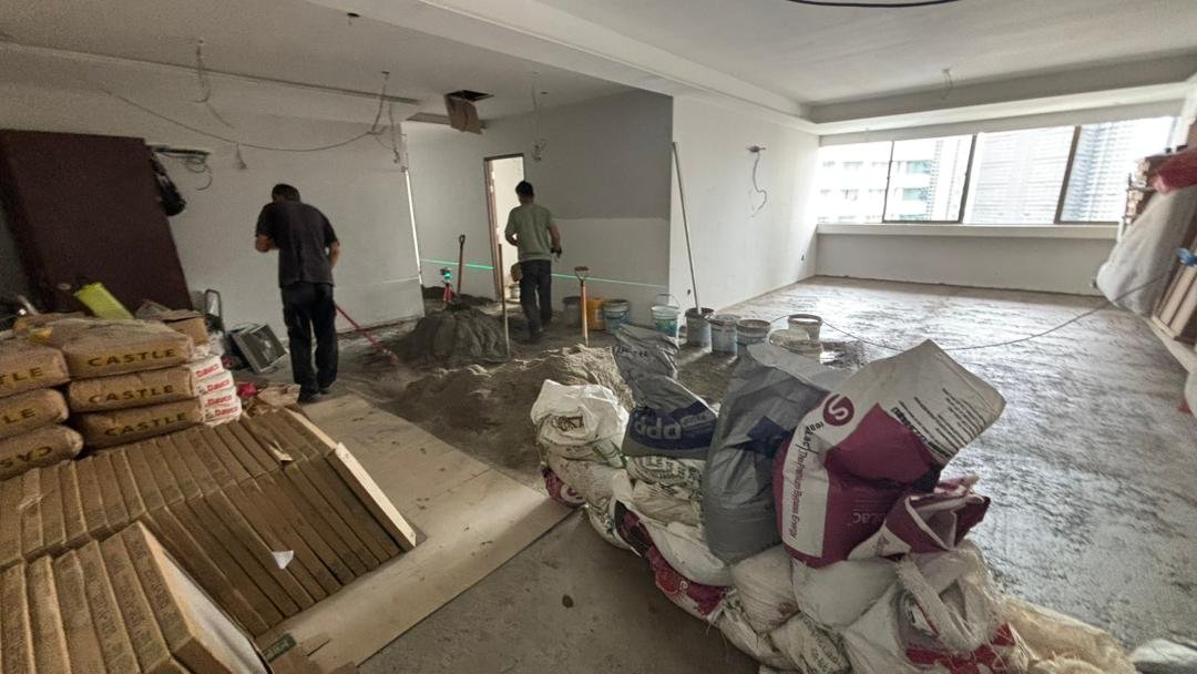 Interior of a room under construction with two workers. Piles of cement bags, construction materials, and tools are visible, with some partially on the floor and some stacked. The room has large windows and an unfinished floor.