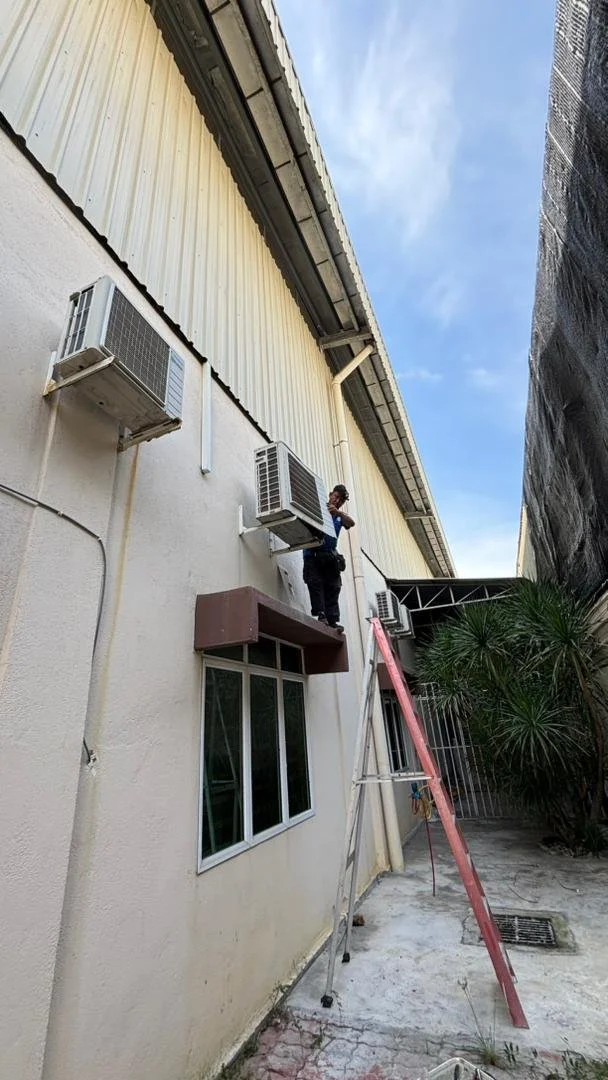 A person standing on an air conditioning unit outside a building, working on the unit, with a ladder positioned against the wall in an alleyway surrounded by a palm tree and construction materials.