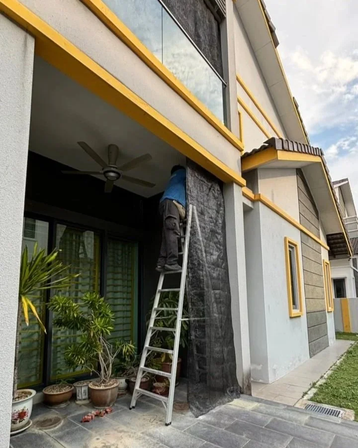 A person in a blue shirt and gray pants is standing on a ladder, working on the exterior of a modern house. They are near a sliding glass door, with potted plants and outdoor tiles in the foreground.