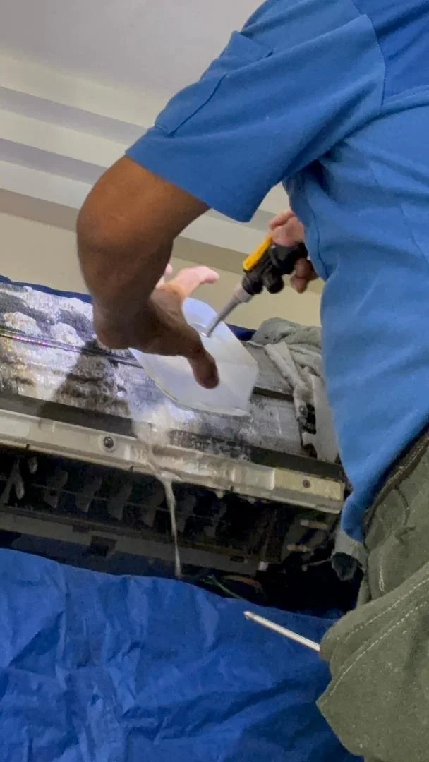 Technician repairing an air conditioning unit using a screwdriver and a syringe-based tool, with water dripping from the unit.