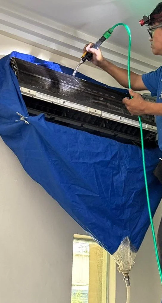 A technician cleaning an air conditioning unit mounted high on a wall using a spray gun connected to hoses.