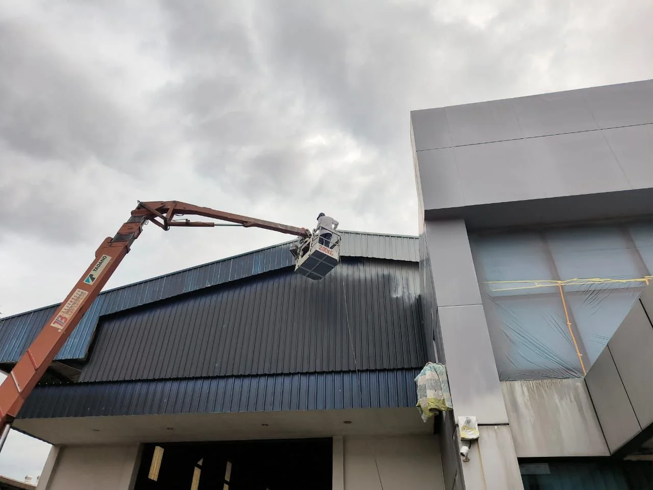 A worker on a lift near the roof of a building, with gray clouds overhead and the building's modern facade.
