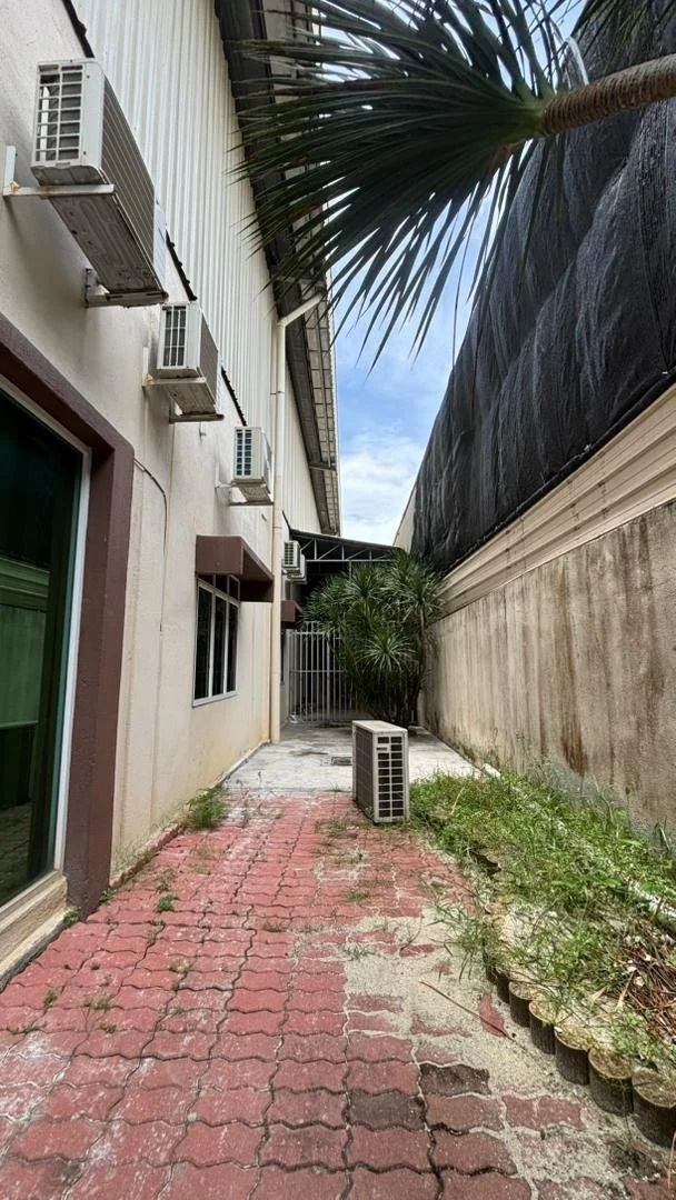 A narrow outdoor alleyway with a red brick paver walkway, a beige building on the left with multiple air conditioning units attached, a large black tarp or covering on the right side wall, and green plants including a small palm tree and weeds growin