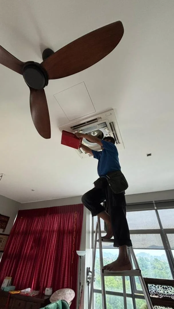 A person standing on a ladder cleaning or servicing a ceiling-mounted air conditioning unit in a living room with a ceiling fan, red curtains, and large windows.