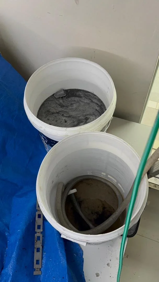 Two white buckets containing mixed materials, with cleaning or construction tools nearby, on a blue surface next to a wall.