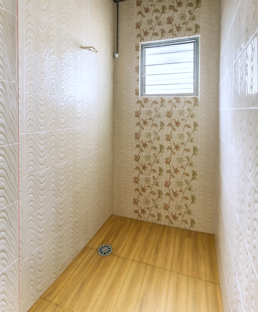 Empty shower stall with tiled walls featuring floral and wave patterns, small window at the top, and wooden floor with drain.