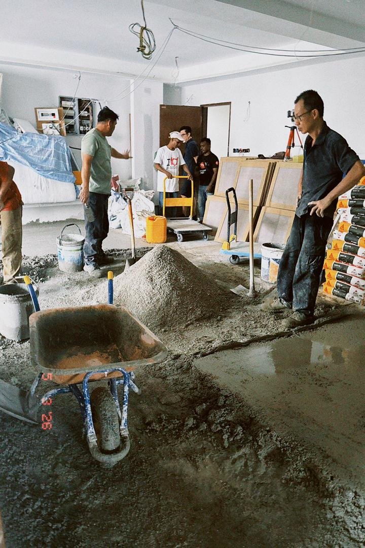 Construction workers are working on a flooring project indoors, with some smoothing cement and others observing. Construction materials and tools are scattered around.