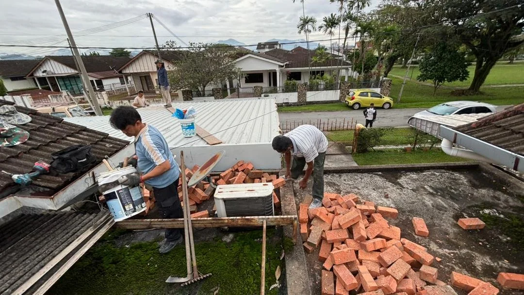 Two men working on a rooftop, with bricks and tools around, preparing for construction or repair amidst a residential neighborhood with trees, cars, and houses.