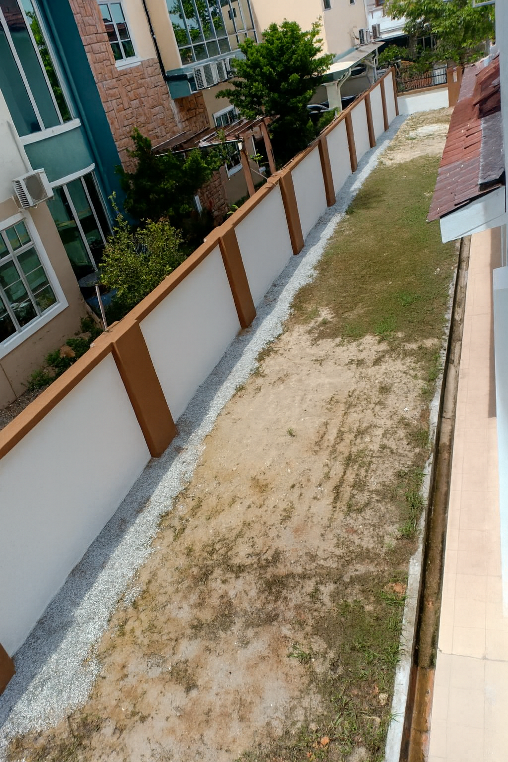 A narrow backyard with a white fence, some green grass, and patches of dirt. On the left, there are residential buildings with windows, air conditioning units, and trees.
