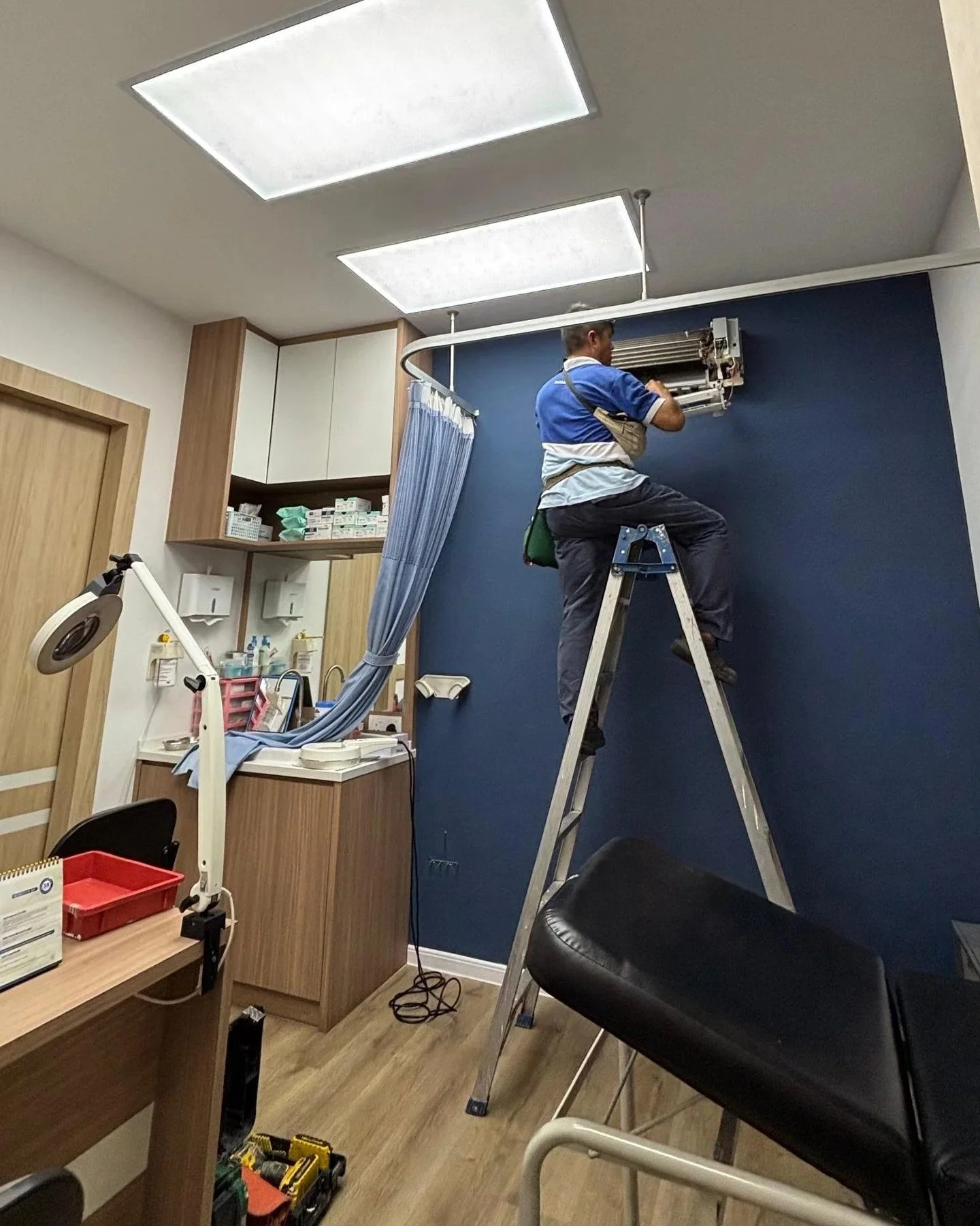 A technician standing on a ladder repairing an air conditioning unit in a medical clinic or office room.