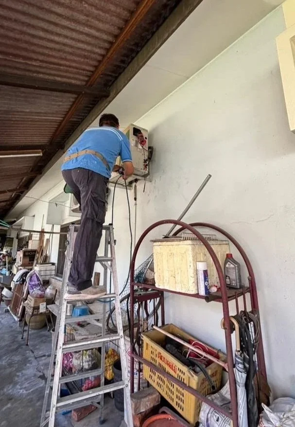 A man standing on a ladder working on an electrical panel mounted on a white wall indoors, with various tools and items on shelves nearby.
