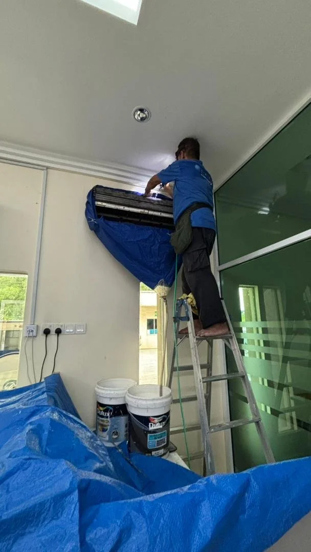 A worker standing on a ladder installing or repairing an air conditioning unit on a wall inside a building.