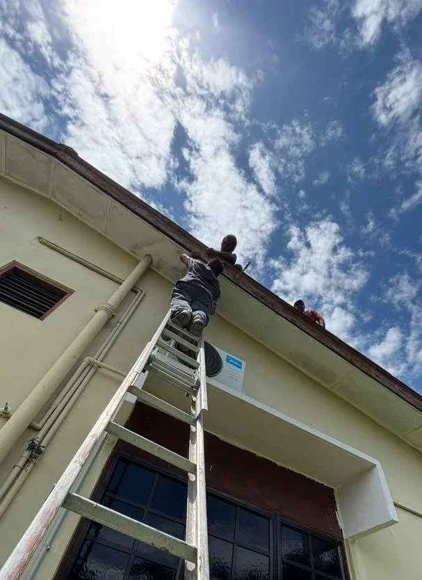 Person on a ladder fixing or inspecting the building's exterior near the roof, with another person watching from the roof, under a partly cloudy sky.