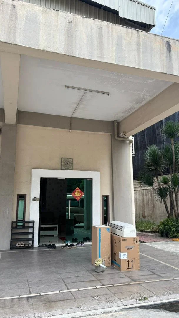 Residential entrance porch with several boxes and an air conditioning unit on the ground, shoes and slippers near the door, with a red and gold decoration hanging inside, and some greenery on the right side.